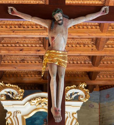 Zdjęcie nr 5: Łuk tęczowy o półkolistym wykroju, na profilowanej belce zdobionej pośrodku ryzowaną rozetą ustawiona grupa Ukrzyżowania. W centrum krucyfiks, po prawej św. Jan Ewangelista, po lewej Matka Boska i św. Maria Magdalena obejmująca krzyż. Chrystus przybity do krzyża trzema gwoździami, sylwetka w delikatnym zwisie, z głową pochyloną delikatnie w dół, o sztywnym układzie sylwetki z zaznaczonymi śladami męki. Perizonium w formie tkaniny przewiązanej na prawym biodrze ze zwisem wzdłuż prawego uda. Twarz podłużna, okolona zarostem; włosy długie spływające na prawe ramię w silnie skręcone pukle, na głowie zielona korona cierniowa złożona z dwóch przeplatających się gałązek. Nad głową Chrystusa biały titulus z napisem: „I(ESUS) N(AZARENUS) R(EX) I(UDAEORUM)”. Krzyż prosty, gładki o ramionach zakończonych gzymsikami. Po lewej stronie krucyfiksu rzeźba św. Marii Magdaleny ukazana w pozycji klęczącej, pochylonej do przodu, obejmująca dłońmi krzyż. Święta jest ubrana w długą, srebrzoną suknię, odsłaniającą nagie ramiona, przewiązaną w talii. Twarz szeroka, delikatnie otwarte usta, małe oczy i długi nos. Głowę okalają długie, ciemne włosy, spływające na plecy. Za nią ustawiona jest rzeźba Matki Boskiej w delikatnym kontrapoście, z prawą nogą ugiętą w kolanie, ze splecionymi na wysokości piersi rękami. Maria jest ubrana w długą, złoconą suknię oraz złocony płaszcz założony na głowę i spływający wzdłuż sylwetki. Twarz szeroka, z wyraźnie zaznaczonym podbródkiem, wzrok skierowany na krucyfiks, zmarszczone brwi. Po prawej stronie rzeźba św. Jana Ewangelisty, ukazanego w postawie stojącej, zwróconego w trzech czwartych w prawo, z rękami splecionymi na wysokości pasa. Twarz owalna, małe usta, długi i wąski nos, wzrok skierowany ku górze, zmarszczone brwi. Włosy ciemne, bujne, spływające na plecy. Święty ubrany jest w długą, złoconą suknię z długimi rękawami. Rzeźby ustawione są na niskich, zielonych cokolikach. Polichromia w odsłoniętych partiach ciała naturalistyczna, detale złocone i srebrzone. 

