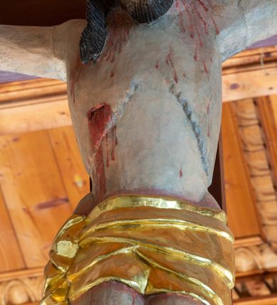 Zdjęcie nr 8: Łuk tęczowy o półkolistym wykroju, na profilowanej belce zdobionej pośrodku ryzowaną rozetą ustawiona grupa Ukrzyżowania. W centrum krucyfiks, po prawej św. Jan Ewangelista, po lewej Matka Boska i św. Maria Magdalena obejmująca krzyż. Chrystus przybity do krzyża trzema gwoździami, sylwetka w delikatnym zwisie, z głową pochyloną delikatnie w dół, o sztywnym układzie sylwetki z zaznaczonymi śladami męki. Perizonium w formie tkaniny przewiązanej na prawym biodrze ze zwisem wzdłuż prawego uda. Twarz podłużna, okolona zarostem; włosy długie spływające na prawe ramię w silnie skręcone pukle, na głowie zielona korona cierniowa złożona z dwóch przeplatających się gałązek. Nad głową Chrystusa biały titulus z napisem: „I(ESUS) N(AZARENUS) R(EX) I(UDAEORUM)”. Krzyż prosty, gładki o ramionach zakończonych gzymsikami. Po lewej stronie krucyfiksu rzeźba św. Marii Magdaleny ukazana w pozycji klęczącej, pochylonej do przodu, obejmująca dłońmi krzyż. Święta jest ubrana w długą, srebrzoną suknię, odsłaniającą nagie ramiona, przewiązaną w talii. Twarz szeroka, delikatnie otwarte usta, małe oczy i długi nos. Głowę okalają długie, ciemne włosy, spływające na plecy. Za nią ustawiona jest rzeźba Matki Boskiej w delikatnym kontrapoście, z prawą nogą ugiętą w kolanie, ze splecionymi na wysokości piersi rękami. Maria jest ubrana w długą, złoconą suknię oraz złocony płaszcz założony na głowę i spływający wzdłuż sylwetki. Twarz szeroka, z wyraźnie zaznaczonym podbródkiem, wzrok skierowany na krucyfiks, zmarszczone brwi. Po prawej stronie rzeźba św. Jana Ewangelisty, ukazanego w postawie stojącej, zwróconego w trzech czwartych w prawo, z rękami splecionymi na wysokości pasa. Twarz owalna, małe usta, długi i wąski nos, wzrok skierowany ku górze, zmarszczone brwi. Włosy ciemne, bujne, spływające na plecy. Święty ubrany jest w długą, złoconą suknię z długimi rękawami. Rzeźby ustawione są na niskich, zielonych cokolikach. Polichromia w odsłoniętych partiach ciała naturalistyczna, detale złocone i srebrzone. 
