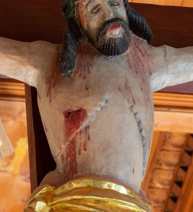 Zdjęcie nr 7: Łuk tęczowy o półkolistym wykroju, na profilowanej belce zdobionej pośrodku ryzowaną rozetą ustawiona grupa Ukrzyżowania. W centrum krucyfiks, po prawej św. Jan Ewangelista, po lewej Matka Boska i św. Maria Magdalena obejmująca krzyż. Chrystus przybity do krzyża trzema gwoździami, sylwetka w delikatnym zwisie, z głową pochyloną delikatnie w dół, o sztywnym układzie sylwetki z zaznaczonymi śladami męki. Perizonium w formie tkaniny przewiązanej na prawym biodrze ze zwisem wzdłuż prawego uda. Twarz podłużna, okolona zarostem; włosy długie spływające na prawe ramię w silnie skręcone pukle, na głowie zielona korona cierniowa złożona z dwóch przeplatających się gałązek. Nad głową Chrystusa biały titulus z napisem: „I(ESUS) N(AZARENUS) R(EX) I(UDAEORUM)”. Krzyż prosty, gładki o ramionach zakończonych gzymsikami. Po lewej stronie krucyfiksu rzeźba św. Marii Magdaleny ukazana w pozycji klęczącej, pochylonej do przodu, obejmująca dłońmi krzyż. Święta jest ubrana w długą, srebrzoną suknię, odsłaniającą nagie ramiona, przewiązaną w talii. Twarz szeroka, delikatnie otwarte usta, małe oczy i długi nos. Głowę okalają długie, ciemne włosy, spływające na plecy. Za nią ustawiona jest rzeźba Matki Boskiej w delikatnym kontrapoście, z prawą nogą ugiętą w kolanie, ze splecionymi na wysokości piersi rękami. Maria jest ubrana w długą, złoconą suknię oraz złocony płaszcz założony na głowę i spływający wzdłuż sylwetki. Twarz szeroka, z wyraźnie zaznaczonym podbródkiem, wzrok skierowany na krucyfiks, zmarszczone brwi. Po prawej stronie rzeźba św. Jana Ewangelisty, ukazanego w postawie stojącej, zwróconego w trzech czwartych w prawo, z rękami splecionymi na wysokości pasa. Twarz owalna, małe usta, długi i wąski nos, wzrok skierowany ku górze, zmarszczone brwi. Włosy ciemne, bujne, spływające na plecy. Święty ubrany jest w długą, złoconą suknię z długimi rękawami. Rzeźby ustawione są na niskich, zielonych cokolikach. Polichromia w odsłoniętych partiach ciała naturalistyczna, detale złocone i srebrzone. 
