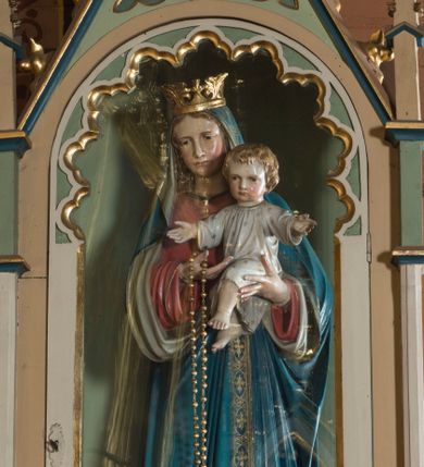 Zdjęcie nr 1: Pełnoplastyczna rzeźba przedstawia Matkę Boską z Dzieciątkiem w całej postaci stojącą na globie ziemskim oplecionym przez węża. Madonna z nieznacznie pochyloną głową w prawo została ujęta w kontrapoście, trzymając Dzieciątko na wysokości piersi lewą ręką, prawą zaś na nie wskazuje. Na prawej ręce ma zawieszony duży, złoty różaniec. Pociągłą twarz Matki Boskiej charakteryzują jasna karnacja z różowymi policzkami, długi, prosty nos, wąskie usta. Jej zamyślone spojrzenie skierowane jest ku dołowi. Twarz otaczają długie, jasnobrązowe włosy opadające na ramiona widoczne spod maforionu. Madonna ubrana jest w niebieski maforion podbity jasnozgniłą zielenią, lamowany złotą, ornamentalną taśmą dekorowaną stylizowanymi krzyżykami. Jego obie poły podciągnięte są na wysokość jej piersi. Spod maforionu widoczna jest czerwona tunika dekorowana przy dekolcie, rękawach i u spodu złotą tasiemką. Tunika odsłania fragmenty stóp w sandałach. Maria ma na głowie złoconą koronę o sterczynach utworzonych ze stylizowanych liści akantu. Dzieciątko zostało ukazane frontalnie z rozłożonymi rękami na wysokości piersi oraz z nieznacznie skrzyżowanymi, bosymi nóżkami. Mały Jezus ma owalną twarz o jasnej karnacji z wysokim czołem, rumiane, pucułowate policzki, szeroko otwarte oczy, wydatny nos i usta. Jego głowę otaczają jasnobrązowe, krótkie włosy. Jezus ubrany jest w jasnoszarą tunikę ze złotym wykończeniem, dekorowaną złotymi gwiazdkami. Pod stopami Marii widnieje do połowy ciemnoniebieski glob ze złotymi gwiazdkami opleciony przez węża, którego depcze lewą stopą Maria. Z przodu globu umieszczono krótką banderolę z łacińskim napisem ”Ave Maria”. Rzeźba znajduje się w przeszklonej witrynie w kształcie neogotyckiej kapliczki z wnęką. Kapliczka uzyskała kształt wydłużonego prostopadłościanu nakrytego dachem siodłowym. Od frontu zaopatrzono ją w półokrągłe, przeszklone drzwi w górnej części dekorowane wewnętrzną arkaturą. Boki flankują smukłe filarki wspierające się na wysokich cokolikach podtrzymujące smukłe imposty z pinaklami zdobionymi czołgankami i zwieńczonymi kwiatonami. Złocone czołganki i kwiaton wieńczący kapliczkę widoczny jest na trójkątnym przyczółku. Jego pole udekorowano płyciną ze złoconym czteroliściem wpisanym w koło otoczone przez trzy trójliście.   