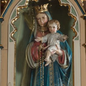 Zdjęcie nr 1: Pełnoplastyczna rzeźba przedstawia Matkę Boską z Dzieciątkiem w całej postaci stojącą na globie ziemskim oplecionym przez węża. Madonna z nieznacznie pochyloną głową w prawo została ujęta w kontrapoście, trzymając Dzieciątko na wysokości piersi lewą ręką, prawą zaś na nie wskazuje. Na prawej ręce ma zawieszony duży, złoty różaniec. Pociągłą twarz Matki Boskiej charakteryzują jasna karnacja z różowymi policzkami, długi, prosty nos, wąskie usta. Jej zamyślone spojrzenie skierowane jest ku dołowi. Twarz otaczają długie, jasnobrązowe włosy opadające na ramiona widoczne spod maforionu. Madonna ubrana jest w niebieski maforion podbity jasnozgniłą zielenią, lamowany złotą, ornamentalną taśmą dekorowaną stylizowanymi krzyżykami. Jego obie poły podciągnięte są na wysokość jej piersi. Spod maforionu widoczna jest czerwona tunika dekorowana przy dekolcie, rękawach i u spodu złotą tasiemką. Tunika odsłania fragmenty stóp w sandałach. Maria ma na głowie złoconą koronę o sterczynach utworzonych ze stylizowanych liści akantu. Dzieciątko zostało ukazane frontalnie z rozłożonymi rękami na wysokości piersi oraz z nieznacznie skrzyżowanymi, bosymi nóżkami. Mały Jezus ma owalną twarz o jasnej karnacji z wysokim czołem, rumiane, pucułowate policzki, szeroko otwarte oczy, wydatny nos i usta. Jego głowę otaczają jasnobrązowe, krótkie włosy. Jezus ubrany jest w jasnoszarą tunikę ze złotym wykończeniem, dekorowaną złotymi gwiazdkami. Pod stopami Marii widnieje do połowy ciemnoniebieski glob ze złotymi gwiazdkami opleciony przez węża, którego depcze lewą stopą Maria. Z przodu globu umieszczono krótką banderolę z łacińskim napisem ”Ave Maria”. Rzeźba znajduje się w przeszklonej witrynie w kształcie neogotyckiej kapliczki z wnęką. Kapliczka uzyskała kształt wydłużonego prostopadłościanu nakrytego dachem siodłowym. Od frontu zaopatrzono ją w półokrągłe, przeszklone drzwi w górnej części dekorowane wewnętrzną arkaturą. Boki flankują smukłe filarki wspierające się na wysokich cokolikach podtrzymujące smukłe imposty z pinaklami zdobionymi czołgankami i zwieńczonymi kwiatonami. Złocone czołganki i kwiaton wieńczący kapliczkę widoczny jest na trójkątnym przyczółku. Jego pole udekorowano płyciną ze złoconym czteroliściem wpisanym w koło otoczone przez trzy trójliście.   