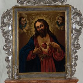 Zdjęcie nr 1: Feretron procesyjny tworzą dwa malowane na płótnie olejne obrazy ujęte w srebrzone ramy, których krawędzie wyznaczają profilowane i złocone listwy, ustawione na prostopadłościennym postumencie. Po bokach ram znajdują się ażurowe, srebrzone uszaki powstałe ze stylizowanych wici akantowych. Zwieńczenie feretronu utworzono z owalnego kartusza otoczonego ażurową, srebrzoną stylizowaną wicią akantową. Na awersie feretronu umieszczono przedstawienie Najświętszego Serca Pana Jezusa, a na rewersie św. Izydora. Postument został zaopatrzony w parę otworów do włożenia drążków umożliwiających noszenie feretronu. Podstawę i zwieńczenie postumentu dekoruje profilowana listwa, natomiast boki jasna brązowo-ochrowa marmoryzacja. Na awersie feretronu wyobrażono w trzech czwartych stojącego Chrystusa ukazującego swoje serce. Zbawiciel ma lekko pochyloną głowę w prawo, obiema zaś przebitymi dłońmi rozsuwa tunikę na piersi, odsłaniając przebite serce. Serce otaczają  złociste promienie. Z rany oraz u góry serca widać wydobywające się płomienie. Twarz Chrystusa jest pociągła. Otaczają ją długie brązowe włosy opadające puklami na ramiona oraz brązowa broda na dole nieznacznie rozdwojona. Twarz Zbawiciela charakteryzuje się wysokim czołem, długim, wąskim nosem i wydatnymi ustami. Chrystus szeroko otwartymi niebieskimi oczami wpatruje się w widza. Jego głowę otaczają złociste promienie. Ubrany jest w luźną, czerwoną tunikę przewiązaną w pasie jasnoochrową szarfą i narzucony na ramiona niebieski płaszcz, którego prawa poła przerzucona jest przez prawe przedramię. Zbawiciel został ukazany na jasnoochrowym tle. Po obu stronach jego głowy na jasnych obłokach widać dwie główki uskrzydlonych aniołków. Mają okrągłe głowy o pucołowatych policzkach z wydatnymi noskami i ustami. Unoszą się na niebieskich skrzydełkach. Na rewersie feretronu umieszczono przedstawienie św. Izydora. Święty został ukazany podczas modlitwy na pierwszym planie, klęcząc przed fragmentem bryły kościoła z półokrągłym oknem o mocno wyartykułowanym glifie. Górną część świątyni okrywają obłoki, z których wyłania się Madonna z Dzieciątkiem. Izydor ukazany został w całej postaci zwrócony w trzech czwartych w prawo z modlitewnie złożonymi rękami na wysokości piersi. O lewe przedramię ma oparty drewniany rydel u dołu okuty metalem. Jego głowa otoczona świetlistym nimbem jest lekko pochylona w prawo. Ma owalną twarz o wyrazistych rysach z dużym nosem i mięsistymi wargami oraz smutnie patrzącymi oczami. Otaczają ją krótkie brązowe włosy odsłaniające lewe ucho oraz krótki zarost. Ubrany jest w białą koszulę z aksamitką pod szyją, zapiętą zieloną kamizelkę oraz rozpięty niebieski surdut z rzędem złotych guzików i czerwone spodnie. Na nogach ma brązowe buty z wysokimi cholewami. Przy jego kolanach pod niewielkim drzewkiem leży brązowy kapelusz z szerokim rondem. W lewym rogu obrazu wyobrażono siedzącą na obłokach Madonnę, ukazaną w trzech czwartych w prawo. Przed sobą obiema rękami trzyma na kolanach Dzieciątko. Maria ma owalną twarz o jasnej karnacji z wydatnym nosem i ustami. Jej  głowę okoloną świetlistym nimbem otaczają długie brązowe włosy opadające na ramiona. Wokół wydekoltowanej szyi widać białą chustę skrzyżowaną na piersi. Madonna ubrana jest w czerwoną suknię przewiązaną w talii i niebieski płaszcz narzucony na ramiona, którego poły okrywają jej kolana. Mały Jezus przedstawiony jest w trzech czwartych w prawo. W lewej ręce trzyma błękitny glob, a prawą unosi w geście błogosławieństwa. Ma owalną twarz o jasnej karnacji z wydatnym nosem i ustami oraz wysokim czołem. Głowę okalają krótkie włosy i otacza świetlisty nimb. Zbawiciel ubrany jest w jasną tunikę odsłaniającą fragment stóp. Na drugim planie za modlącym się Izydorem wyobrażono dwa epizody z jego legendy. Z lewej strony pod ścianą kościoła widać Izydora nauczającego pięcioosobową grupę siedzących przed nim na ziemi ludzi. Święty obiema rękami trzyma książkę. Ubrany jest w kapelusz, białą koszulę, niebieski surdut, czerwone spodnie i buty z wysokimi cholewami. Dwóch ze słuchaczy siedzących z prawej strony ubranych jest w jasne szaty i zawoje na głowach. Natomiast trójka pozostałych to mężczyzna w kapeluszu z kosturem w lewej ręce odziany w łachmany, a także dwie postacie ukazane od tyłu ubrane w szaty z kapturami na głowach. Z tyłu z prawej strony Izydora widać anioła orzącego ziemię pługiem ciągnionym przez woła. Anioł z rozpostartymi skrzydłami w nieznacznym rozkroku ujęty został w trzech czwartych w lewo. Ubrany jest w tunikę sięgającą kolan z rozcięciem u dołu z przodu oraz sandały wiązane pod kolanami. Za nim w oddali widać klęczącego Izydora przed drewnianym krzyżem zatkniętym na skalnym wzgórku przy którym rosną trzy drzewa ułożone w szeregu. Tłem dla całego przedstawienia jest ciemne burzowe niebo rozjaśnione przy linii horyzontu. W lewym, dolnym rogu obrazu widać jasny napis „S(ANCTUS) ISIDORUS / AGRICOLA, HIS”.

