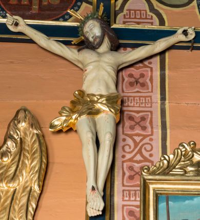 Zdjęcie nr 1: Na niebieskim, drewnianym krzyżu o ramionach wykończonych profilowanymi, złoconymi listwami zawieszono pełnoplastyczną rzeźbę ukrzyżowanego Chrystusa. Ponad jego głową przytwierdzono złocony titulus w formie krótkiej, wygiętej, podłużnej banderoli z inskrypcją "IN / RI". Ukrzyżowany Chrystus o jasnej karnacji ciała ma ukośnie wyciągnięte, sztywne ręce  o przybitych do krzyża dłoniach oraz założoną nogę prawą na lewą, przytwierdzone jednym gwoździem. Jego ciało jest szczupłe. Ukrzyżowany ma zapadniętą klatkę piersiową, podkreślone żebra i obojczyk. Głowa Jezusa w zielonej koronie cierniowej opada na prawe ramię. Twarz otacza krótki brązowy zarost oraz długie brązowe włosy przykrywające kark i opadające na prawe ramię. Zbawiciel ma zamknięte oczy, prosty nos i wąskie usta. Na czole, szyi, ramionach, przy przebitym boku, dłoniach i stopach oraz na nogach widoczne są stróżki krwi. W partii bioder ciało Chrystusa okrywa przewiązane, złocone perizonium przechodzące w rozwiany feston przy prawym boku.