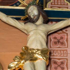Zdjęcie nr 1: Na niebieskim, drewnianym krzyżu o ramionach wykończonych profilowanymi, złoconymi listwami zawieszono pełnoplastyczną rzeźbę ukrzyżowanego Chrystusa. Ponad jego głową przytwierdzono złocony titulus w formie krótkiej, wygiętej, podłużnej banderoli z inskrypcją "IN / RI". Ukrzyżowany Chrystus o jasnej karnacji ciała ma ukośnie wyciągnięte, sztywne ręce  o przybitych do krzyża dłoniach oraz założoną nogę prawą na lewą, przytwierdzone jednym gwoździem. Jego ciało jest szczupłe. Ukrzyżowany ma zapadniętą klatkę piersiową, podkreślone żebra i obojczyk. Głowa Jezusa w zielonej koronie cierniowej opada na prawe ramię. Twarz otacza krótki brązowy zarost oraz długie brązowe włosy przykrywające kark i opadające na prawe ramię. Zbawiciel ma zamknięte oczy, prosty nos i wąskie usta. Na czole, szyi, ramionach, przy przebitym boku, dłoniach i stopach oraz na nogach widoczne są stróżki krwi. W partii bioder ciało Chrystusa okrywa przewiązane, złocone perizonium przechodzące w rozwiany feston przy prawym boku.