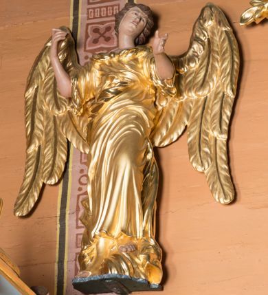 Zdjęcie nr 1: Pełnoplastyczna wolnostojąca figura przedstawia w całej postaci uskrzydlonego anioła. Został on ujęty frontalnie w kontrapoście z głową w trzech czwartych zwróconą w prawo w lekkim skłonie. Stoi on na złocistym obłoku mając lewą nogę lekko zgiętą w kolanie i wspartą o obłok. Anioł obie ręce ma uniesione ku górze na wysokości piersi. Układ palców prawej ręki wskazuje, iż mógł w niej coś trzymać, natomiast lewa dłoń ujęta jest w geście wskazującym. Postać charakteryzuje pociągła, młodzieńcza twarz o jasnej karnacji z nieznacznie zmarszczonymi brwiami, prostym nosem i półotwartymi ustami. Jego głowę okalają modelowane brązowe włosy sięgające karku. Złocone skrzydła anioła są rozpostarta z charakterystycznym mocnym wycięciem i mięsistym modelunkiem piór ułożonych wertykalnie. Anioł ubrany jest w sięgającą ziemie złoconą tunikę odsłaniającą bose stopy obute w wysokie sandały. Ekspresyjnie udrapowana tunika ma krótkie rękawy sięgające łokci.