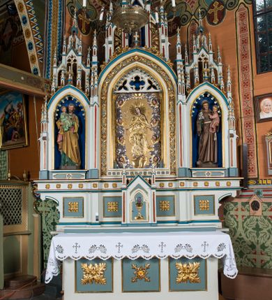 Zdjęcie nr 1: Ołtarz otrzymał formę drewnianej, przyściennej struktury architektonicznej ze zwieńczeniem o trójosiowym, jednokondygnacyjnym założeniu. Nastawa prezentuje się jako struktura polichromowana w odcieniach jasnej seledynowej zieleni, rozbielonego granatu oraz czerwieni. Partie ornamentów zostały wyzłocone. Drewniana mensa z portatylem wspiera się na prostopadłościennym stipesie ołtarzowym. Jego przód zdobią trzy czworoboczne płaszczyzny zbliżone do kwadratu ujęte profilowanymi, złoconymi listwami. W ich centrum umieszczono płaskorzeźbione, złocone czteroliście w diagonalnym układzie. Dwa w skrajnych płycinach utworzono ze stylizowanych liści akantu, natomiast środkowy przyjął formę geometryczno-roślinną. Retabulum ustawiono na wysokiej podstawie w kształcie nawiązującym do wydłużonego trapezu z wklęsłymi bokami, wspierającej się na krótszym boku. Podstawę zamyka u góry łamana, profilowana listwa. Jej lico artykułowane jest czterema zbliżonymi do kwadratu płycinami obramionymi złotymi, profilowanymi listwami. Ich środek zdobią takie same stylizowane, płaskorzeźbione i złocone czteroliście o diagonalnym układzie, wpisane w kwadrat. Na środku mensy, na tle podstawy, ustawiono czworoboczne tabernakulum w formie kapliczki z dwuspadowym dachem flankowanej dwiema sterczynami z pinaklami, wyrastającymi z wydłużonych prostopadłościennych, wąskich przypór. Drzwiczki tabernakulum zdobi motyw płaskorzeźbionego kielicha ze srebrzoną hostią wpisaną w stylizowaną ostrołukową arkadę z noskami. Na podstawie nastawy wspiera się niski cokół zamknięty u góry fryzem z rytmicznie rozmieszczonymi, niewielkimi, płaskorzeźbionymi i złoconymi czterolistnymi rozetkami. Z cokołu wyrastają trzy ostrołukowe arkady o profilowanych krawędziach, w górnych częściach z wpisaną arkaturą, ozdobione płaskorzeźbionymi, złoconymi czołgankami, zwieńczone pinaklami. Arkada środkowa jest wyższa od flankujących ją arkad bocznych. Zdobi ją po bokach płaskorzeźbione obramienie z motywem złoconej wici roślinno-ornamentalnej. Ostrołukowe zamknięcie arkady wypełniono ażurowym maswerkiem z motywem czteroliścia wpisanego w koło, ujętego dwoma trójliściami. Wszystkie trzy arkady flankują smukłe sterczyny wyrastające z prostopadłościennych, wąskich przypór ustawionych na niskich cokolikach. Sterczyny przechodzą w pinakle zwieńczone kwiatonami. Dekorują je płaskorzeźbione i złocone czołganki. W przypadku arkad bocznych pomiędzy cokolikami wprowadzono podłużne płyciny ze stylizowanymi maswerkami, natomiast w arkadzie centralnej umieszczono niewielkie, diagonalne czteroliście. W polu środkowej arkady prezentowany jest obraz Matki Boskiej Niepokalanie Poczętej w płaskorzeźbionej, złoconej, drewnianej sukience wotywnej, a na zasuwach wizerunek Przemienienia Pańskiego i św. Walentego. W arkadach bocznych retabulum ustawiono z lewej strony pełnoplastyczną, polichromowaną rzeźbę z masy gipsowej św. Józefa z Dzieciątkiem, a z prawej św. Antoniego Padewskiego, także z Dzieciątkiem Jezus. Zwieńczenie nastawy zostało również ukształtowane architektonicznie. Tworzą je trzy ażurowe kapliczki z pinaklami ustawione na osiach retabulum. W kapliczce środkowej umieszczono pełnoplastyczną, polichromowaną figurę św. Franciszka z Asyżu z masy gipsowej. 