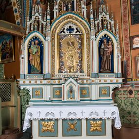 Zdjęcie nr 1: Ołtarz otrzymał formę drewnianej, przyściennej struktury architektonicznej ze zwieńczeniem o trójosiowym, jednokondygnacyjnym założeniu. Nastawa prezentuje się jako struktura polichromowana w odcieniach jasnej seledynowej zieleni, rozbielonego granatu oraz czerwieni. Partie ornamentów zostały wyzłocone. Drewniana mensa z portatylem wspiera się na prostopadłościennym stipesie ołtarzowym. Jego przód zdobią trzy czworoboczne płaszczyzny zbliżone do kwadratu ujęte profilowanymi, złoconymi listwami. W ich centrum umieszczono płaskorzeźbione, złocone czteroliście w diagonalnym układzie. Dwa w skrajnych płycinach utworzono ze stylizowanych liści akantu, natomiast środkowy przyjął formę geometryczno-roślinną. Retabulum ustawiono na wysokiej podstawie w kształcie nawiązującym do wydłużonego trapezu z wklęsłymi bokami, wspierającej się na krótszym boku. Podstawę zamyka u góry łamana, profilowana listwa. Jej lico artykułowane jest czterema zbliżonymi do kwadratu płycinami obramionymi złotymi, profilowanymi listwami. Ich środek zdobią takie same stylizowane, płaskorzeźbione i złocone czteroliście o diagonalnym układzie, wpisane w kwadrat. Na środku mensy, na tle podstawy, ustawiono czworoboczne tabernakulum w formie kapliczki z dwuspadowym dachem flankowanej dwiema sterczynami z pinaklami, wyrastającymi z wydłużonych prostopadłościennych, wąskich przypór. Drzwiczki tabernakulum zdobi motyw płaskorzeźbionego kielicha ze srebrzoną hostią wpisaną w stylizowaną ostrołukową arkadę z noskami. Na podstawie nastawy wspiera się niski cokół zamknięty u góry fryzem z rytmicznie rozmieszczonymi, niewielkimi, płaskorzeźbionymi i złoconymi czterolistnymi rozetkami. Z cokołu wyrastają trzy ostrołukowe arkady o profilowanych krawędziach, w górnych częściach z wpisaną arkaturą, ozdobione płaskorzeźbionymi, złoconymi czołgankami, zwieńczone pinaklami. Arkada środkowa jest wyższa od flankujących ją arkad bocznych. Zdobi ją po bokach płaskorzeźbione obramienie z motywem złoconej wici roślinno-ornamentalnej. Ostrołukowe zamknięcie arkady wypełniono ażurowym maswerkiem z motywem czteroliścia wpisanego w koło, ujętego dwoma trójliściami. Wszystkie trzy arkady flankują smukłe sterczyny wyrastające z prostopadłościennych, wąskich przypór ustawionych na niskich cokolikach. Sterczyny przechodzą w pinakle zwieńczone kwiatonami. Dekorują je płaskorzeźbione i złocone czołganki. W przypadku arkad bocznych pomiędzy cokolikami wprowadzono podłużne płyciny ze stylizowanymi maswerkami, natomiast w arkadzie centralnej umieszczono niewielkie, diagonalne czteroliście. W polu środkowej arkady prezentowany jest obraz Matki Boskiej Niepokalanie Poczętej w płaskorzeźbionej, złoconej, drewnianej sukience wotywnej, a na zasuwach wizerunek Przemienienia Pańskiego i św. Walentego. W arkadach bocznych retabulum ustawiono z lewej strony pełnoplastyczną, polichromowaną rzeźbę z masy gipsowej św. Józefa z Dzieciątkiem, a z prawej św. Antoniego Padewskiego, także z Dzieciątkiem Jezus. Zwieńczenie nastawy zostało również ukształtowane architektonicznie. Tworzą je trzy ażurowe kapliczki z pinaklami ustawione na osiach retabulum. W kapliczce środkowej umieszczono pełnoplastyczną, polichromowaną figurę św. Franciszka z Asyżu z masy gipsowej. 