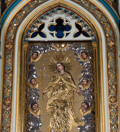 Zdjęcie nr 2: Ołtarz otrzymał formę drewnianej, przyściennej struktury architektonicznej ze zwieńczeniem o trójosiowym, jednokondygnacyjnym założeniu. Nastawa prezentuje się jako struktura polichromowana w odcieniach jasnej seledynowej zieleni, rozbielonego granatu oraz czerwieni. Partie ornamentów zostały wyzłocone. Drewniana mensa z portatylem wspiera się na prostopadłościennym stipesie ołtarzowym. Jego przód zdobią trzy czworoboczne płaszczyzny zbliżone do kwadratu ujęte profilowanymi, złoconymi listwami. W ich centrum umieszczono płaskorzeźbione, złocone czteroliście w diagonalnym układzie. Dwa w skrajnych płycinach utworzono ze stylizowanych liści akantu, natomiast środkowy przyjął formę geometryczno-roślinną. Retabulum ustawiono na wysokiej podstawie w kształcie nawiązującym do wydłużonego trapezu z wklęsłymi bokami, wspierającej się na krótszym boku. Podstawę zamyka u góry łamana, profilowana listwa. Jej lico artykułowane jest czterema zbliżonymi do kwadratu płycinami obramionymi złotymi, profilowanymi listwami. Ich środek zdobią takie same stylizowane, płaskorzeźbione i złocone czteroliście o diagonalnym układzie, wpisane w kwadrat. Na środku mensy, na tle podstawy, ustawiono czworoboczne tabernakulum w formie kapliczki z dwuspadowym dachem flankowanej dwiema sterczynami z pinaklami, wyrastającymi z wydłużonych prostopadłościennych, wąskich przypór. Drzwiczki tabernakulum zdobi motyw płaskorzeźbionego kielicha ze srebrzoną hostią wpisaną w stylizowaną ostrołukową arkadę z noskami. Na podstawie nastawy wspiera się niski cokół zamknięty u góry fryzem z rytmicznie rozmieszczonymi, niewielkimi, płaskorzeźbionymi i złoconymi czterolistnymi rozetkami. Z cokołu wyrastają trzy ostrołukowe arkady o profilowanych krawędziach, w górnych częściach z wpisaną arkaturą, ozdobione płaskorzeźbionymi, złoconymi czołgankami, zwieńczone pinaklami. Arkada środkowa jest wyższa od flankujących ją arkad bocznych. Zdobi ją po bokach płaskorzeźbione obramienie z motywem złoconej wici roślinno-ornamentalnej. Ostrołukowe zamknięcie arkady wypełniono ażurowym maswerkiem z motywem czteroliścia wpisanego w koło, ujętego dwoma trójliściami. Wszystkie trzy arkady flankują smukłe sterczyny wyrastające z prostopadłościennych, wąskich przypór ustawionych na niskich cokolikach. Sterczyny przechodzą w pinakle zwieńczone kwiatonami. Dekorują je płaskorzeźbione i złocone czołganki. W przypadku arkad bocznych pomiędzy cokolikami wprowadzono podłużne płyciny ze stylizowanymi maswerkami, natomiast w arkadzie centralnej umieszczono niewielkie, diagonalne czteroliście. W polu środkowej arkady prezentowany jest obraz Matki Boskiej Niepokalanie Poczętej w płaskorzeźbionej, złoconej, drewnianej sukience wotywnej, a na zasuwach wizerunek Przemienienia Pańskiego i św. Walentego. W arkadach bocznych retabulum ustawiono z lewej strony pełnoplastyczną, polichromowaną rzeźbę z masy gipsowej św. Józefa z Dzieciątkiem, a z prawej św. Antoniego Padewskiego, także z Dzieciątkiem Jezus. Zwieńczenie nastawy zostało również ukształtowane architektonicznie. Tworzą je trzy ażurowe kapliczki z pinaklami ustawione na osiach retabulum. W kapliczce środkowej umieszczono pełnoplastyczną, polichromowaną figurę św. Franciszka z Asyżu z masy gipsowej. 