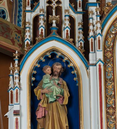 Zdjęcie nr 9: Ołtarz otrzymał formę drewnianej, przyściennej struktury architektonicznej ze zwieńczeniem o trójosiowym, jednokondygnacyjnym założeniu. Nastawa prezentuje się jako struktura polichromowana w odcieniach jasnej seledynowej zieleni, rozbielonego granatu oraz czerwieni. Partie ornamentów zostały wyzłocone. Drewniana mensa z portatylem wspiera się na prostopadłościennym stipesie ołtarzowym. Jego przód zdobią trzy czworoboczne płaszczyzny zbliżone do kwadratu ujęte profilowanymi, złoconymi listwami. W ich centrum umieszczono płaskorzeźbione, złocone czteroliście w diagonalnym układzie. Dwa w skrajnych płycinach utworzono ze stylizowanych liści akantu, natomiast środkowy przyjął formę geometryczno-roślinną. Retabulum ustawiono na wysokiej podstawie w kształcie nawiązującym do wydłużonego trapezu z wklęsłymi bokami, wspierającej się na krótszym boku. Podstawę zamyka u góry łamana, profilowana listwa. Jej lico artykułowane jest czterema zbliżonymi do kwadratu płycinami obramionymi złotymi, profilowanymi listwami. Ich środek zdobią takie same stylizowane, płaskorzeźbione i złocone czteroliście o diagonalnym układzie, wpisane w kwadrat. Na środku mensy, na tle podstawy, ustawiono czworoboczne tabernakulum w formie kapliczki z dwuspadowym dachem flankowanej dwiema sterczynami z pinaklami, wyrastającymi z wydłużonych prostopadłościennych, wąskich przypór. Drzwiczki tabernakulum zdobi motyw płaskorzeźbionego kielicha ze srebrzoną hostią wpisaną w stylizowaną ostrołukową arkadę z noskami. Na podstawie nastawy wspiera się niski cokół zamknięty u góry fryzem z rytmicznie rozmieszczonymi, niewielkimi, płaskorzeźbionymi i złoconymi czterolistnymi rozetkami. Z cokołu wyrastają trzy ostrołukowe arkady o profilowanych krawędziach, w górnych częściach z wpisaną arkaturą, ozdobione płaskorzeźbionymi, złoconymi czołgankami, zwieńczone pinaklami. Arkada środkowa jest wyższa od flankujących ją arkad bocznych. Zdobi ją po bokach płaskorzeźbione obramienie z motywem złoconej wici roślinno-ornamentalnej. Ostrołukowe zamknięcie arkady wypełniono ażurowym maswerkiem z motywem czteroliścia wpisanego w koło, ujętego dwoma trójliściami. Wszystkie trzy arkady flankują smukłe sterczyny wyrastające z prostopadłościennych, wąskich przypór ustawionych na niskich cokolikach. Sterczyny przechodzą w pinakle zwieńczone kwiatonami. Dekorują je płaskorzeźbione i złocone czołganki. W przypadku arkad bocznych pomiędzy cokolikami wprowadzono podłużne płyciny ze stylizowanymi maswerkami, natomiast w arkadzie centralnej umieszczono niewielkie, diagonalne czteroliście. W polu środkowej arkady prezentowany jest obraz Matki Boskiej Niepokalanie Poczętej w płaskorzeźbionej, złoconej, drewnianej sukience wotywnej, a na zasuwach wizerunek Przemienienia Pańskiego i św. Walentego. W arkadach bocznych retabulum ustawiono z lewej strony pełnoplastyczną, polichromowaną rzeźbę z masy gipsowej św. Józefa z Dzieciątkiem, a z prawej św. Antoniego Padewskiego, także z Dzieciątkiem Jezus. Zwieńczenie nastawy zostało również ukształtowane architektonicznie. Tworzą je trzy ażurowe kapliczki z pinaklami ustawione na osiach retabulum. W kapliczce środkowej umieszczono pełnoplastyczną, polichromowaną figurę św. Franciszka z Asyżu z masy gipsowej. 