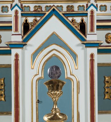 Zdjęcie nr 11: Ołtarz otrzymał formę drewnianej, przyściennej struktury architektonicznej ze zwieńczeniem o trójosiowym, jednokondygnacyjnym założeniu. Nastawa prezentuje się jako struktura polichromowana w odcieniach jasnej seledynowej zieleni, rozbielonego granatu oraz czerwieni. Partie ornamentów zostały wyzłocone. Drewniana mensa z portatylem wspiera się na prostopadłościennym stipesie ołtarzowym. Jego przód zdobią trzy czworoboczne płaszczyzny zbliżone do kwadratu ujęte profilowanymi, złoconymi listwami. W ich centrum umieszczono płaskorzeźbione, złocone czteroliście w diagonalnym układzie. Dwa w skrajnych płycinach utworzono ze stylizowanych liści akantu, natomiast środkowy przyjął formę geometryczno-roślinną. Retabulum ustawiono na wysokiej podstawie w kształcie nawiązującym do wydłużonego trapezu z wklęsłymi bokami, wspierającej się na krótszym boku. Podstawę zamyka u góry łamana, profilowana listwa. Jej lico artykułowane jest czterema zbliżonymi do kwadratu płycinami obramionymi złotymi, profilowanymi listwami. Ich środek zdobią takie same stylizowane, płaskorzeźbione i złocone czteroliście o diagonalnym układzie, wpisane w kwadrat. Na środku mensy, na tle podstawy, ustawiono czworoboczne tabernakulum w formie kapliczki z dwuspadowym dachem flankowanej dwiema sterczynami z pinaklami, wyrastającymi z wydłużonych prostopadłościennych, wąskich przypór. Drzwiczki tabernakulum zdobi motyw płaskorzeźbionego kielicha ze srebrzoną hostią wpisaną w stylizowaną ostrołukową arkadę z noskami. Na podstawie nastawy wspiera się niski cokół zamknięty u góry fryzem z rytmicznie rozmieszczonymi, niewielkimi, płaskorzeźbionymi i złoconymi czterolistnymi rozetkami. Z cokołu wyrastają trzy ostrołukowe arkady o profilowanych krawędziach, w górnych częściach z wpisaną arkaturą, ozdobione płaskorzeźbionymi, złoconymi czołgankami, zwieńczone pinaklami. Arkada środkowa jest wyższa od flankujących ją arkad bocznych. Zdobi ją po bokach płaskorzeźbione obramienie z motywem złoconej wici roślinno-ornamentalnej. Ostrołukowe zamknięcie arkady wypełniono ażurowym maswerkiem z motywem czteroliścia wpisanego w koło, ujętego dwoma trójliściami. Wszystkie trzy arkady flankują smukłe sterczyny wyrastające z prostopadłościennych, wąskich przypór ustawionych na niskich cokolikach. Sterczyny przechodzą w pinakle zwieńczone kwiatonami. Dekorują je płaskorzeźbione i złocone czołganki. W przypadku arkad bocznych pomiędzy cokolikami wprowadzono podłużne płyciny ze stylizowanymi maswerkami, natomiast w arkadzie centralnej umieszczono niewielkie, diagonalne czteroliście. W polu środkowej arkady prezentowany jest obraz Matki Boskiej Niepokalanie Poczętej w płaskorzeźbionej, złoconej, drewnianej sukience wotywnej, a na zasuwach wizerunek Przemienienia Pańskiego i św. Walentego. W arkadach bocznych retabulum ustawiono z lewej strony pełnoplastyczną, polichromowaną rzeźbę z masy gipsowej św. Józefa z Dzieciątkiem, a z prawej św. Antoniego Padewskiego, także z Dzieciątkiem Jezus. Zwieńczenie nastawy zostało również ukształtowane architektonicznie. Tworzą je trzy ażurowe kapliczki z pinaklami ustawione na osiach retabulum. W kapliczce środkowej umieszczono pełnoplastyczną, polichromowaną figurę św. Franciszka z Asyżu z masy gipsowej. 