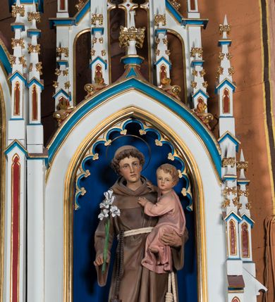 Zdjęcie nr 6: Ołtarz otrzymał formę drewnianej, przyściennej struktury architektonicznej ze zwieńczeniem o trójosiowym, jednokondygnacyjnym założeniu. Nastawa prezentuje się jako struktura polichromowana w odcieniach jasnej seledynowej zieleni, rozbielonego granatu oraz czerwieni. Partie ornamentów zostały wyzłocone. Drewniana mensa z portatylem wspiera się na prostopadłościennym stipesie ołtarzowym. Jego przód zdobią trzy czworoboczne płaszczyzny zbliżone do kwadratu ujęte profilowanymi, złoconymi listwami. W ich centrum umieszczono płaskorzeźbione, złocone czteroliście w diagonalnym układzie. Dwa w skrajnych płycinach utworzono ze stylizowanych liści akantu, natomiast środkowy przyjął formę geometryczno-roślinną. Retabulum ustawiono na wysokiej podstawie w kształcie nawiązującym do wydłużonego trapezu z wklęsłymi bokami, wspierającej się na krótszym boku. Podstawę zamyka u góry łamana, profilowana listwa. Jej lico artykułowane jest czterema zbliżonymi do kwadratu płycinami obramionymi złotymi, profilowanymi listwami. Ich środek zdobią takie same stylizowane, płaskorzeźbione i złocone czteroliście o diagonalnym układzie, wpisane w kwadrat. Na środku mensy, na tle podstawy, ustawiono czworoboczne tabernakulum w formie kapliczki z dwuspadowym dachem flankowanej dwiema sterczynami z pinaklami, wyrastającymi z wydłużonych prostopadłościennych, wąskich przypór. Drzwiczki tabernakulum zdobi motyw płaskorzeźbionego kielicha ze srebrzoną hostią wpisaną w stylizowaną ostrołukową arkadę z noskami. Na podstawie nastawy wspiera się niski cokół zamknięty u góry fryzem z rytmicznie rozmieszczonymi, niewielkimi, płaskorzeźbionymi i złoconymi czterolistnymi rozetkami. Z cokołu wyrastają trzy ostrołukowe arkady o profilowanych krawędziach, w górnych częściach z wpisaną arkaturą, ozdobione płaskorzeźbionymi, złoconymi czołgankami, zwieńczone pinaklami. Arkada środkowa jest wyższa od flankujących ją arkad bocznych. Zdobi ją po bokach płaskorzeźbione obramienie z motywem złoconej wici roślinno-ornamentalnej. Ostrołukowe zamknięcie arkady wypełniono ażurowym maswerkiem z motywem czteroliścia wpisanego w koło, ujętego dwoma trójliściami. Wszystkie trzy arkady flankują smukłe sterczyny wyrastające z prostopadłościennych, wąskich przypór ustawionych na niskich cokolikach. Sterczyny przechodzą w pinakle zwieńczone kwiatonami. Dekorują je płaskorzeźbione i złocone czołganki. W przypadku arkad bocznych pomiędzy cokolikami wprowadzono podłużne płyciny ze stylizowanymi maswerkami, natomiast w arkadzie centralnej umieszczono niewielkie, diagonalne czteroliście. W polu środkowej arkady prezentowany jest obraz Matki Boskiej Niepokalanie Poczętej w płaskorzeźbionej, złoconej, drewnianej sukience wotywnej, a na zasuwach wizerunek Przemienienia Pańskiego i św. Walentego. W arkadach bocznych retabulum ustawiono z lewej strony pełnoplastyczną, polichromowaną rzeźbę z masy gipsowej św. Józefa z Dzieciątkiem, a z prawej św. Antoniego Padewskiego, także z Dzieciątkiem Jezus. Zwieńczenie nastawy zostało również ukształtowane architektonicznie. Tworzą je trzy ażurowe kapliczki z pinaklami ustawione na osiach retabulum. W kapliczce środkowej umieszczono pełnoplastyczną, polichromowaną figurę św. Franciszka z Asyżu z masy gipsowej. 