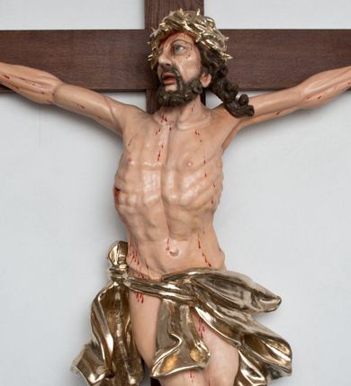 Zdjęcie nr 1: Postać ukrzyżowanego Chrystusa została przymocowana do brązowego, drewnianego krzyża za pomocą trzech gwoździ. Pełna ekspresji rzeźba ukazuje Chrystusa z głową uniesioną ponad rozpięte ramiona ułożone w lekkim zwisie. Jezus w esowatym ujęciu został ukazany jako żywy. Jego nogi ugięte są nieznacznie w kolanach. Prawa stopa założona jest na lewą. Głowa Zbawiciela zwrócona jest w prawą stronę. Ciało posiada malowaną jasną karnację. Korpus rzeźbiony jest płasko z charakterystycznym zaznaczeniem żeber oraz z nienaturalnym duktem długiego mostka. Z prawej strony zaznaczono krwawiącą ranę w boku. Zbawiciel ma pociągłą, szczupłą twarz, okoloną zmierzwionymi, brązowymi włosami nieznacznie rozwianymi z lewej strony oraz zarostem. Cechą charakterystyczną twarzy są szeroko otwarte, niebieskie oczy, duży, mięsisty nos oraz wydatne usta nieznacznie rozchylone. Jezus na głowie ma srebrzoną koronę cierniową, spod której spływa krew. Krew została zaznaczona przy gwoździach oraz na ciele. Biodra Zbawiciela okrywa rozwiane srebrzone, perizonium o ekspresyjnym układzie draperii, zawinięte po obu stronach za podwójny sznur widoczny na prawym biodrze. Ponad głową Chrystusa zamocowano na krzyżu titulus w formie srebrzonej, rozwianej banderoli z napisem „IN/RI”.