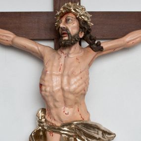 Zdjęcie nr 1: Postać ukrzyżowanego Chrystusa została przymocowana do brązowego, drewnianego krzyża za pomocą trzech gwoździ. Pełna ekspresji rzeźba ukazuje Chrystusa z głową uniesioną ponad rozpięte ramiona ułożone w lekkim zwisie. Jezus w esowatym ujęciu został ukazany jako żywy. Jego nogi ugięte są nieznacznie w kolanach. Prawa stopa założona jest na lewą. Głowa Zbawiciela zwrócona jest w prawą stronę. Ciało posiada malowaną jasną karnację. Korpus rzeźbiony jest płasko z charakterystycznym zaznaczeniem żeber oraz z nienaturalnym duktem długiego mostka. Z prawej strony zaznaczono krwawiącą ranę w boku. Zbawiciel ma pociągłą, szczupłą twarz, okoloną zmierzwionymi, brązowymi włosami nieznacznie rozwianymi z lewej strony oraz zarostem. Cechą charakterystyczną twarzy są szeroko otwarte, niebieskie oczy, duży, mięsisty nos oraz wydatne usta nieznacznie rozchylone. Jezus na głowie ma srebrzoną koronę cierniową, spod której spływa krew. Krew została zaznaczona przy gwoździach oraz na ciele. Biodra Zbawiciela okrywa rozwiane srebrzone, perizonium o ekspresyjnym układzie draperii, zawinięte po obu stronach za podwójny sznur widoczny na prawym biodrze. Ponad głową Chrystusa zamocowano na krzyżu titulus w formie srebrzonej, rozwianej banderoli z napisem „IN/RI”.
