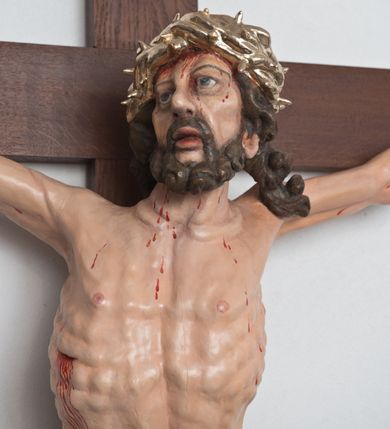 Zdjęcie nr 2: Postać ukrzyżowanego Chrystusa została przymocowana do brązowego, drewnianego krzyża za pomocą trzech gwoździ. Pełna ekspresji rzeźba ukazuje Chrystusa z głową uniesioną ponad rozpięte ramiona ułożone w lekkim zwisie. Jezus w esowatym ujęciu został ukazany jako żywy. Jego nogi ugięte są nieznacznie w kolanach. Prawa stopa założona jest na lewą. Głowa Zbawiciela zwrócona jest w prawą stronę. Ciało posiada malowaną jasną karnację. Korpus rzeźbiony jest płasko z charakterystycznym zaznaczeniem żeber oraz z nienaturalnym duktem długiego mostka. Z prawej strony zaznaczono krwawiącą ranę w boku. Zbawiciel ma pociągłą, szczupłą twarz, okoloną zmierzwionymi, brązowymi włosami nieznacznie rozwianymi z lewej strony oraz zarostem. Cechą charakterystyczną twarzy są szeroko otwarte, niebieskie oczy, duży, mięsisty nos oraz wydatne usta nieznacznie rozchylone. Jezus na głowie ma srebrzoną koronę cierniową, spod której spływa krew. Krew została zaznaczona przy gwoździach oraz na ciele. Biodra Zbawiciela okrywa rozwiane srebrzone, perizonium o ekspresyjnym układzie draperii, zawinięte po obu stronach za podwójny sznur widoczny na prawym biodrze. Ponad głową Chrystusa zamocowano na krzyżu titulus w formie srebrzonej, rozwianej banderoli z napisem „IN/RI”.