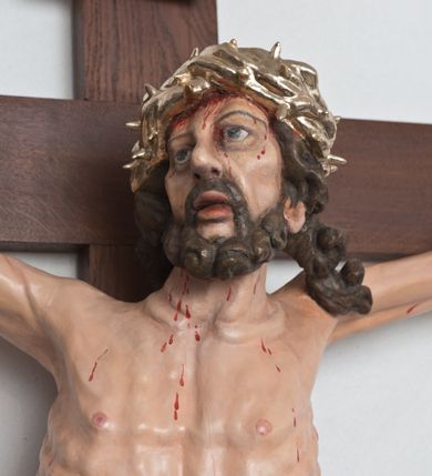Zdjęcie nr 3: Postać ukrzyżowanego Chrystusa została przymocowana do brązowego, drewnianego krzyża za pomocą trzech gwoździ. Pełna ekspresji rzeźba ukazuje Chrystusa z głową uniesioną ponad rozpięte ramiona ułożone w lekkim zwisie. Jezus w esowatym ujęciu został ukazany jako żywy. Jego nogi ugięte są nieznacznie w kolanach. Prawa stopa założona jest na lewą. Głowa Zbawiciela zwrócona jest w prawą stronę. Ciało posiada malowaną jasną karnację. Korpus rzeźbiony jest płasko z charakterystycznym zaznaczeniem żeber oraz z nienaturalnym duktem długiego mostka. Z prawej strony zaznaczono krwawiącą ranę w boku. Zbawiciel ma pociągłą, szczupłą twarz, okoloną zmierzwionymi, brązowymi włosami nieznacznie rozwianymi z lewej strony oraz zarostem. Cechą charakterystyczną twarzy są szeroko otwarte, niebieskie oczy, duży, mięsisty nos oraz wydatne usta nieznacznie rozchylone. Jezus na głowie ma srebrzoną koronę cierniową, spod której spływa krew. Krew została zaznaczona przy gwoździach oraz na ciele. Biodra Zbawiciela okrywa rozwiane srebrzone, perizonium o ekspresyjnym układzie draperii, zawinięte po obu stronach za podwójny sznur widoczny na prawym biodrze. Ponad głową Chrystusa zamocowano na krzyżu titulus w formie srebrzonej, rozwianej banderoli z napisem „IN/RI”.