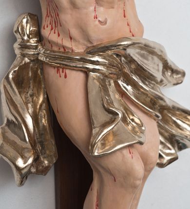 Zdjęcie nr 4: Postać ukrzyżowanego Chrystusa została przymocowana do brązowego, drewnianego krzyża za pomocą trzech gwoździ. Pełna ekspresji rzeźba ukazuje Chrystusa z głową uniesioną ponad rozpięte ramiona ułożone w lekkim zwisie. Jezus w esowatym ujęciu został ukazany jako żywy. Jego nogi ugięte są nieznacznie w kolanach. Prawa stopa założona jest na lewą. Głowa Zbawiciela zwrócona jest w prawą stronę. Ciało posiada malowaną jasną karnację. Korpus rzeźbiony jest płasko z charakterystycznym zaznaczeniem żeber oraz z nienaturalnym duktem długiego mostka. Z prawej strony zaznaczono krwawiącą ranę w boku. Zbawiciel ma pociągłą, szczupłą twarz, okoloną zmierzwionymi, brązowymi włosami nieznacznie rozwianymi z lewej strony oraz zarostem. Cechą charakterystyczną twarzy są szeroko otwarte, niebieskie oczy, duży, mięsisty nos oraz wydatne usta nieznacznie rozchylone. Jezus na głowie ma srebrzoną koronę cierniową, spod której spływa krew. Krew została zaznaczona przy gwoździach oraz na ciele. Biodra Zbawiciela okrywa rozwiane srebrzone, perizonium o ekspresyjnym układzie draperii, zawinięte po obu stronach za podwójny sznur widoczny na prawym biodrze. Ponad głową Chrystusa zamocowano na krzyżu titulus w formie srebrzonej, rozwianej banderoli z napisem „IN/RI”.