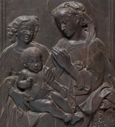 Zdjęcie nr 1: Relief powstał w wyniku odlewu. Uzyskał on kształt stojącego prostokąta ujętego profilowaną ramą. W centrum kompozycji wyobrażono adorującą Matkę Boską Dzieciątko Jezus. Siedzącą w półpostaci Marię ukazano w trzech czwartych w lewo. Jest to młoda kobieta, która na wysokości piersi trzyma złożone modlitewnie dłonie. Na jej kolanach siedzi mały Jezus podtrzymywany przez stojącego za nim młodzieńca. Twarz Madonny charakteryzują miękkie rysy, wysokie czoło, prosty nos i wydatne usta. Spojrzenie jej półprzymkniętych oczu skierowane jest na Jezusa. Falujące włosy otaczające głowę i odsłaniające lewe ucho, ukryte są pod delikatną chustą zawiązaną na głowie, której fragment opada na lewe ramię. Maria ubrana jest w suknię obszytą pod szyją haftowaną taśmą z widoczną na przedzie główką serafina oraz luźny płaszcz opadający miękkimi fałdami na ramię i widoczny fragment oparcia krzesła nożycowego. Nad głową Marii widoczny jest zaznaczony za pomocą lini nimb talerzowy.
Dzieciątko Jezus zostało ukazane frontalnie w mocnym kontrapoście. Jezus prawą rękę zgiętą w łokciu trzyma wysuniętą przed siebie. Natomiast w lewej ułożonej wzdłuż tułowia ma jabłko. Jego owalną twarz charakteryzuje wysokie czoło, wydatny nos i usta. Spojrzenie jego otwartych oczu skierowane jest na widza. Głowę Dzieciątka okalają krótkie włosy odsłaniające uszy. Nagość małego Jezusa okrywa jedynie w części korpusu fragment tkaniny przewiązany w pasie. Dlatego widoczne jest prawe ramię oraz bose nogi od pasa w dół. Siedzącego na kolanach Marii Jezusa obiema rękami podtrzymuje młodzieniec stojący z tyłu z lewej strony kompozycji. Został on ukazany frontalnie z głową ujętą w trzech czwartych w prawo. Ma owalną twarz z wysokim czołem, wydatnym nosem i ustami. Jego głowę otaczają lekko rozwiane kędzierzawe włosy, odsłaniające końcówkę prawego ucha. Ubrany jest w tunikę lamowaną pod szyją i narzucony na ramiona płaszcz. W prawym dolnym rogu kompozycji widoczne jest wolutowe oparcie krzesła nożycowego, zdobione perełkowaniem i stylizowanym liściem akantu.