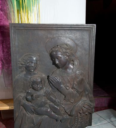 Zdjęcie nr 3: Relief powstał w wyniku odlewu. Uzyskał on kształt stojącego prostokąta ujętego profilowaną ramą. W centrum kompozycji wyobrażono adorującą Matkę Boską Dzieciątko Jezus. Siedzącą w półpostaci Marię ukazano w trzech czwartych w lewo. Jest to młoda kobieta, która na wysokości piersi trzyma złożone modlitewnie dłonie. Na jej kolanach siedzi mały Jezus podtrzymywany przez stojącego za nim młodzieńca. Twarz Madonny charakteryzują miękkie rysy, wysokie czoło, prosty nos i wydatne usta. Spojrzenie jej półprzymkniętych oczu skierowane jest na Jezusa. Falujące włosy otaczające głowę i odsłaniające lewe ucho, ukryte są pod delikatną chustą zawiązaną na głowie, której fragment opada na lewe ramię. Maria ubrana jest w suknię obszytą pod szyją haftowaną taśmą z widoczną na przedzie główką serafina oraz luźny płaszcz opadający miękkimi fałdami na ramię i widoczny fragment oparcia krzesła nożycowego. Nad głową Marii widoczny jest zaznaczony za pomocą lini nimb talerzowy.
Dzieciątko Jezus zostało ukazane frontalnie w mocnym kontrapoście. Jezus prawą rękę zgiętą w łokciu trzyma wysuniętą przed siebie. Natomiast w lewej ułożonej wzdłuż tułowia ma jabłko. Jego owalną twarz charakteryzuje wysokie czoło, wydatny nos i usta. Spojrzenie jego otwartych oczu skierowane jest na widza. Głowę Dzieciątka okalają krótkie włosy odsłaniające uszy. Nagość małego Jezusa okrywa jedynie w części korpusu fragment tkaniny przewiązany w pasie. Dlatego widoczne jest prawe ramię oraz bose nogi od pasa w dół. Siedzącego na kolanach Marii Jezusa obiema rękami podtrzymuje młodzieniec stojący z tyłu z lewej strony kompozycji. Został on ukazany frontalnie z głową ujętą w trzech czwartych w prawo. Ma owalną twarz z wysokim czołem, wydatnym nosem i ustami. Jego głowę otaczają lekko rozwiane kędzierzawe włosy, odsłaniające końcówkę prawego ucha. Ubrany jest w tunikę lamowaną pod szyją i narzucony na ramiona płaszcz. W prawym dolnym rogu kompozycji widoczne jest wolutowe oparcie krzesła nożycowego, zdobione perełkowaniem i stylizowanym liściem akantu.