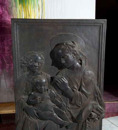 Zdjęcie nr 2: Relief powstał w wyniku odlewu. Uzyskał on kształt stojącego prostokąta ujętego profilowaną ramą. W centrum kompozycji wyobrażono adorującą Matkę Boską Dzieciątko Jezus. Siedzącą w półpostaci Marię ukazano w trzech czwartych w lewo. Jest to młoda kobieta, która na wysokości piersi trzyma złożone modlitewnie dłonie. Na jej kolanach siedzi mały Jezus podtrzymywany przez stojącego za nim młodzieńca. Twarz Madonny charakteryzują miękkie rysy, wysokie czoło, prosty nos i wydatne usta. Spojrzenie jej półprzymkniętych oczu skierowane jest na Jezusa. Falujące włosy otaczające głowę i odsłaniające lewe ucho, ukryte są pod delikatną chustą zawiązaną na głowie, której fragment opada na lewe ramię. Maria ubrana jest w suknię obszytą pod szyją haftowaną taśmą z widoczną na przedzie główką serafina oraz luźny płaszcz opadający miękkimi fałdami na ramię i widoczny fragment oparcia krzesła nożycowego. Nad głową Marii widoczny jest zaznaczony za pomocą lini nimb talerzowy.
Dzieciątko Jezus zostało ukazane frontalnie w mocnym kontrapoście. Jezus prawą rękę zgiętą w łokciu trzyma wysuniętą przed siebie. Natomiast w lewej ułożonej wzdłuż tułowia ma jabłko. Jego owalną twarz charakteryzuje wysokie czoło, wydatny nos i usta. Spojrzenie jego otwartych oczu skierowane jest na widza. Głowę Dzieciątka okalają krótkie włosy odsłaniające uszy. Nagość małego Jezusa okrywa jedynie w części korpusu fragment tkaniny przewiązany w pasie. Dlatego widoczne jest prawe ramię oraz bose nogi od pasa w dół. Siedzącego na kolanach Marii Jezusa obiema rękami podtrzymuje młodzieniec stojący z tyłu z lewej strony kompozycji. Został on ukazany frontalnie z głową ujętą w trzech czwartych w prawo. Ma owalną twarz z wysokim czołem, wydatnym nosem i ustami. Jego głowę otaczają lekko rozwiane kędzierzawe włosy, odsłaniające końcówkę prawego ucha. Ubrany jest w tunikę lamowaną pod szyją i narzucony na ramiona płaszcz. W prawym dolnym rogu kompozycji widoczne jest wolutowe oparcie krzesła nożycowego, zdobione perełkowaniem i stylizowanym liściem akantu.