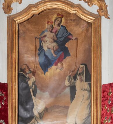 Zdjęcie nr 1: Obraz Matki Boskiej Różańcowej ze świętymi dominikańskimi w kształcie stojącego prostokąta, zamkniętego łukiem wklęsło-wypukłym z uskokami. Kompozycja dwustrefowa, w partii wyższej ukazana jest Matka Boska w pozycji siedzącej na obłokach, frontalnie, z Dzieciątkiem Jezus na prawym boku, lewą ręką podająca różaniec św. Katarzynie. Twarz o delikatnych rysach, oczy przymknięte, wzrok skierowany na św. Katarzynę; jasne i długie włosy. Maria ubrana jest w jasnoróżową suknię z długimi rękawami; błękitny płaszcz otulający całą postać oraz biały welon na głowie. Dzieciątko zwrócone jest trzy czwarte w lewo, ze skrzyżowanymi nogami, prawą ręką podaje różaniec św. Dominikowi, a lewą ma ugiętą w łokciu. Twarz pełna, zarumieniona, włosy jasne i kędzierzawe. Dzieciątko ubrane jest w białą pieluszkę, osłaniającą biodra. Matka Boska i Dzieciątko mają wokół głów koliste nimby, a na głowach zamknięte korony. W dolnej strefie, po lewej stronie obrazu klęczy św. Dominik, zwrócony w lewo, prawą ręką odbierający różaniec od Dzieciątka, a lewą ma złożoną na piersi. Twarz ukazana jest z lewego profilu o rysach twarzy starszego mężczyzny z siwą i krótką brodą, długim nosem, łysiną na głowie i śniadą cerą. Ubrany jest w płaszcz i habit dominikański. Poniżej widoczny jest niebieski glob ziemski oraz głowa psa z pochodnią w pysku. Po prawej stronie klęczy św. Katarzyna ze Sieny, zwrócona trzy czwarte w prawo, która prawą ręką odbiera różaniec od Marii, a lewą unosi ku górze. Twarz owalna z pełnymi ustami, krótkim nosem i dużymi oczami ze wzorkiem skierowanym na Matkę Boską. Ubrana jest w strój dominikański: habit, płaszcz i welon z podwiką. W oddali widoczny jest pejzaż górski i zabudowania. W górnej części kompozycji pochmurne niebo oraz uskrzydlone główki anielskie w narożach oraz jedna przy stopie Matki Boskiej. 
