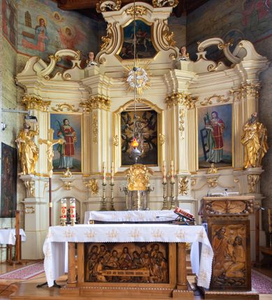 Zdjęcie nr 22: Nastawa ołtarzowa architektoniczna, trójosiowa, jednokondygnacyjna ze zwieńczeniem i bramkami. Ołtarz drewniany, prostopadłościenny; antepedium haftowane. Tabernakulum metalowe, fakturowane, w formie zbliżonej do prostopadłościennej skrzynki, ustawione na niskim cokole, o wypukłej ściance frontowej, flankowane parą kolumn o kompozytowych kapitelach. Drzwiczki w kształcie stojącego prostokąta, zamkniętego łukiem nadwieszonym; w polu kielich z hostią, a po bokach winna winorośl i kłosy zbóż. Powyżej jest tron wystawienia o fantazyjnym kształcie, ujęty po bokach wolutami, na których usadzone są dwa putta. W polu maska lunarna, otoczona promienistą glorią, a w zwieńczeniu muszla palmetowa. Nastawa ustawiona jest na wysokim, zdwojonym cokole, flankowana dwiema parami zdwojonych pilastrów, ustawionych ukośnie. Bramki w kształcie stojących prostokątów, zamkniętych półkoliście, w profilowanej obudowie zwieńczonej wolutowo. Drzwiczki dekorowane są malowanym bukietem kwiatów w wazonie. Kondygnacja o delikatnie wklęsłym przebiegu, ujęta dwiema parami zdwojonych, kanelowanych pilastrów, z których wewnętrzne ustawione są pod kątem. Podpory dźwigają pełne belkowanie, wyłamane nad osiami. Na linii wewnętrznych podpór ustawione są dwa cokoły, na których osadzono dwie główki aniołków, a na linii skrajnych podpór wolutowe przyczółki; z kolei pomiędzy nimi znajdują się fantazyjne woluty. W polu środkowym w ramie o kształcie stojącego prostokąta, zamkniętego łukiem wklęsło-wypukłym z uskokami umieszczony jest obraz Wniebowzięcia Najświętszej Marii Panny, a na zasuwie obraz św. Marcina oraz płótno przedstawiające Chrystusa Ukrzyżowanego. W osiach bocznych obrazy: po lewej św. Szczepan, a po prawej św. Wawrzyniec. Przed skrajnymi podporami ustawione są figury: po lewej św. Piotra, a po prawej św. Pawła. Nad polem głównym zawieszona jest gołębica Ducha Świętego na tle srebrzonych obłoków i promienistej glorii. Zwieńczenie w formie zbliżonej do leżącego prostokąta ujętego spływami wolutowymi, zamknięte profilowanym gzymsem i rocaille’ami. W polu obraz św. Marcina. Struktura polichromowana w kolorze jasnobeżowym, ornamentyka złocona, detale srebrzone.



