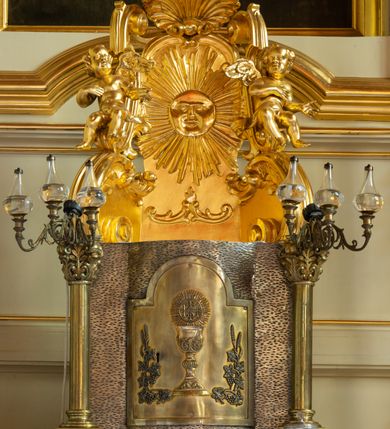 Zdjęcie nr 12: Nastawa ołtarzowa architektoniczna, trójosiowa, jednokondygnacyjna ze zwieńczeniem i bramkami. Ołtarz drewniany, prostopadłościenny; antepedium haftowane. Tabernakulum metalowe, fakturowane, w formie zbliżonej do prostopadłościennej skrzynki, ustawione na niskim cokole, o wypukłej ściance frontowej, flankowane parą kolumn o kompozytowych kapitelach. Drzwiczki w kształcie stojącego prostokąta, zamkniętego łukiem nadwieszonym; w polu kielich z hostią, a po bokach winna winorośl i kłosy zbóż. Powyżej jest tron wystawienia o fantazyjnym kształcie, ujęty po bokach wolutami, na których usadzone są dwa putta. W polu maska lunarna, otoczona promienistą glorią, a w zwieńczeniu muszla palmetowa. Nastawa ustawiona jest na wysokim, zdwojonym cokole, flankowana dwiema parami zdwojonych pilastrów, ustawionych ukośnie. Bramki w kształcie stojących prostokątów, zamkniętych półkoliście, w profilowanej obudowie zwieńczonej wolutowo. Drzwiczki dekorowane są malowanym bukietem kwiatów w wazonie. Kondygnacja o delikatnie wklęsłym przebiegu, ujęta dwiema parami zdwojonych, kanelowanych pilastrów, z których wewnętrzne ustawione są pod kątem. Podpory dźwigają pełne belkowanie, wyłamane nad osiami. Na linii wewnętrznych podpór ustawione są dwa cokoły, na których osadzono dwie główki aniołków, a na linii skrajnych podpór wolutowe przyczółki; z kolei pomiędzy nimi znajdują się fantazyjne woluty. W polu środkowym w ramie o kształcie stojącego prostokąta, zamkniętego łukiem wklęsło-wypukłym z uskokami umieszczony jest obraz Wniebowzięcia Najświętszej Marii Panny, a na zasuwie obraz św. Marcina oraz płótno przedstawiające Chrystusa Ukrzyżowanego. W osiach bocznych obrazy: po lewej św. Szczepan, a po prawej św. Wawrzyniec. Przed skrajnymi podporami ustawione są figury: po lewej św. Piotra, a po prawej św. Pawła. Nad polem głównym zawieszona jest gołębica Ducha Świętego na tle srebrzonych obłoków i promienistej glorii. Zwieńczenie w formie zbliżonej do leżącego prostokąta ujętego spływami wolutowymi, zamknięte profilowanym gzymsem i rocaille’ami. W polu obraz św. Marcina. Struktura polichromowana w kolorze jasnobeżowym, ornamentyka złocona, detale srebrzone.



