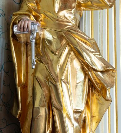 Zdjęcie nr 14: Nastawa ołtarzowa architektoniczna, trójosiowa, jednokondygnacyjna ze zwieńczeniem i bramkami. Ołtarz drewniany, prostopadłościenny; antepedium haftowane. Tabernakulum metalowe, fakturowane, w formie zbliżonej do prostopadłościennej skrzynki, ustawione na niskim cokole, o wypukłej ściance frontowej, flankowane parą kolumn o kompozytowych kapitelach. Drzwiczki w kształcie stojącego prostokąta, zamkniętego łukiem nadwieszonym; w polu kielich z hostią, a po bokach winna winorośl i kłosy zbóż. Powyżej jest tron wystawienia o fantazyjnym kształcie, ujęty po bokach wolutami, na których usadzone są dwa putta. W polu maska lunarna, otoczona promienistą glorią, a w zwieńczeniu muszla palmetowa. Nastawa ustawiona jest na wysokim, zdwojonym cokole, flankowana dwiema parami zdwojonych pilastrów, ustawionych ukośnie. Bramki w kształcie stojących prostokątów, zamkniętych półkoliście, w profilowanej obudowie zwieńczonej wolutowo. Drzwiczki dekorowane są malowanym bukietem kwiatów w wazonie. Kondygnacja o delikatnie wklęsłym przebiegu, ujęta dwiema parami zdwojonych, kanelowanych pilastrów, z których wewnętrzne ustawione są pod kątem. Podpory dźwigają pełne belkowanie, wyłamane nad osiami. Na linii wewnętrznych podpór ustawione są dwa cokoły, na których osadzono dwie główki aniołków, a na linii skrajnych podpór wolutowe przyczółki; z kolei pomiędzy nimi znajdują się fantazyjne woluty. W polu środkowym w ramie o kształcie stojącego prostokąta, zamkniętego łukiem wklęsło-wypukłym z uskokami umieszczony jest obraz Wniebowzięcia Najświętszej Marii Panny, a na zasuwie obraz św. Marcina oraz płótno przedstawiające Chrystusa Ukrzyżowanego. W osiach bocznych obrazy: po lewej św. Szczepan, a po prawej św. Wawrzyniec. Przed skrajnymi podporami ustawione są figury: po lewej św. Piotra, a po prawej św. Pawła. Nad polem głównym zawieszona jest gołębica Ducha Świętego na tle srebrzonych obłoków i promienistej glorii. Zwieńczenie w formie zbliżonej do leżącego prostokąta ujętego spływami wolutowymi, zamknięte profilowanym gzymsem i rocaille’ami. W polu obraz św. Marcina. Struktura polichromowana w kolorze jasnobeżowym, ornamentyka złocona, detale srebrzone.



