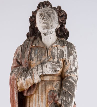 Zdjęcie nr 1: Pełnoplastyczna rzeźba św. Jana Ewangelisty stojącego w kontrapoście, z lewą noga ugiętą w kolanie. Święty jest ukazany frontalnie, prawą rękę składa na piersi, w lewej, opuszczonej wzdłuż ciała trzyma zamkniętą księgę. Głowa odchylona do tyłu, twarz podłużna, z prostym nosem, wąskimi ustami, wydatnym podbródkiem, okolona brązowymi włosami, które opadają na ramiona i plecy. Jan jest ubrany w żółtą, rozciętą pod szyją suknię przepasaną w talii oraz czerwony płaszcz zarzucony na prawe ramię i owijający się wokół lewego biodra, opadający prostymi fałdami w dół. Spod tuniki widoczne bose stopy. 