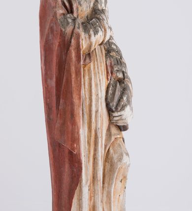 Zdjęcie nr 6: Pełnoplastyczna rzeźba św. Jana Ewangelisty stojącego w kontrapoście, z lewą noga ugiętą w kolanie. Święty jest ukazany frontalnie, prawą rękę składa na piersi, w lewej, opuszczonej wzdłuż ciała trzyma zamkniętą księgę. Głowa odchylona do tyłu, twarz podłużna, z prostym nosem, wąskimi ustami, wydatnym podbródkiem, okolona brązowymi włosami, które opadają na ramiona i plecy. Jan jest ubrany w żółtą, rozciętą pod szyją suknię przepasaną w talii oraz czerwony płaszcz zarzucony na prawe ramię i owijający się wokół lewego biodra, opadający prostymi fałdami w dół. Spod tuniki widoczne bose stopy. 