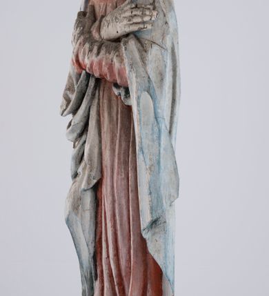 Zdjęcie nr 7: Pełnoplastyczna rzeźba Matki Boskiej Bolesnej ustawionej frontalnie, w lekkim kontrapoście, z prawą nogą ugiętą w kolanie i rękami skrzyżowanymi na piersi. Maria skręca głowę nieznacznie w lewo, spogląda smutno w dół. Na podłużnej twarzy odznaczają się wysokie czoło, duże, migdałowe, półprzymknięte oczy, wąski nos, niewielkie usta oraz szeroki podbródek. Ubrana jest w czerwoną suknię o wąskich, marszczących się równoległymi fałdami rękawach i kołnierzu, spływającą do ziemi podłużnymi fałdami. Na nią nałożony ma jasnoniebieski, zarzucony na głowę płaszcz, który zakrywa plecy i spływa na ramiona. Prawa poła przytrzymana między rękami zawija się przy prawym biodrze łamanymi fałdami. Spod sukni widoczne ciemnobrązowe, szpiczaste buty.