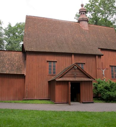 Zdjęcie nr 28: Kościół usytuowany na wzniesieniu, w otoczeniu bogatego drzewostanu. Orientowany, jednonawowy, z zamkniętym trójbocznie prezbiterium i przylegającą od północy zakrystią. Od południa i zachodu do korpusu przylegają kruchty. Nawa na planie prostokąta, szersza od prezbiterium. Wewnątrz pozorne sklepienie kolebkowe, w nawie z płaskimi odcinkami stropowymi po bokach, podpartymi na profilowanych słupach ustawionych na cokołach. Ściany opięte pilastrami naśladującymi formę słupów. Prezbiterium od korpusu oddziela łuk tęczowy, półkolisty, profilowany, ozdobiony ornamentem sznurowym, wsparty na profilowanych kroksztynach dekorowanych drewnianymi rozetami. Od zachodu parapet chóru muzycznego na planie prostokąta oparty na dwóch wielobocznych słupach, front dekorowany motywem w formie powtarzających się zwróconych do siebie par esownic i napisem „MIŁOŚCIĄ WIECZNĄ UMIŁOWAŁEM CIĘ” rozdzielonym wizerunkiem pelikana karmiącego młode własną krwią. Od południa schody prowadzące na chór z analogiczną dekoracją. Fasada prosta, zamknięta daszkiem krytym gontem, zwieńczona trójkątnie. Korpus z trzech stron wzmocniony belkami. Od strony północnej i południowej po dwa okna prostokątne zwieńczone niewielkimi blaszanymi daszkami. Prezbiterium od strony południowej opięte dwoma belkami, z prostokątnym oknem. Na ścianie wschodniej, na zewnątrz, kapliczka z ceramicznym przedstawieniem Matki Boskiej z Dzieciątkiem. W ścianach wschodniej i północnej zakrystii po jednym prostokątnym oknie. Dach dwuspadowy, o jednej kalenicy, od wschodu odzwierciedla układ ścian prezbiterium. Kruchty przykryte dachami dwuspadowymi. Dachy kryte gontem. Na przecięciu korpusu z prezbiterium blaszana, ośmioboczna wieżyczka na sygnaturkę zwieńczona hełmem baniastym ze spłaszczoną kulą z krzyżem. 