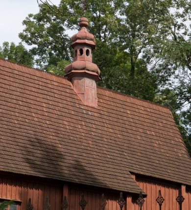 Zdjęcie nr 9: Kościół usytuowany na wzniesieniu, w otoczeniu bogatego drzewostanu. Orientowany, jednonawowy, z zamkniętym trójbocznie prezbiterium i przylegającą od północy zakrystią. Od południa i zachodu do korpusu przylegają kruchty. Nawa na planie prostokąta, szersza od prezbiterium. Wewnątrz pozorne sklepienie kolebkowe, w nawie z płaskimi odcinkami stropowymi po bokach, podpartymi na profilowanych słupach ustawionych na cokołach. Ściany opięte pilastrami naśladującymi formę słupów. Prezbiterium od korpusu oddziela łuk tęczowy, półkolisty, profilowany, ozdobiony ornamentem sznurowym, wsparty na profilowanych kroksztynach dekorowanych drewnianymi rozetami. Od zachodu parapet chóru muzycznego na planie prostokąta oparty na dwóch wielobocznych słupach, front dekorowany motywem w formie powtarzających się zwróconych do siebie par esownic i napisem „MIŁOŚCIĄ WIECZNĄ UMIŁOWAŁEM CIĘ” rozdzielonym wizerunkiem pelikana karmiącego młode własną krwią. Od południa schody prowadzące na chór z analogiczną dekoracją. Fasada prosta, zamknięta daszkiem krytym gontem, zwieńczona trójkątnie. Korpus z trzech stron wzmocniony belkami. Od strony północnej i południowej po dwa okna prostokątne zwieńczone niewielkimi blaszanymi daszkami. Prezbiterium od strony południowej opięte dwoma belkami, z prostokątnym oknem. Na ścianie wschodniej, na zewnątrz, kapliczka z ceramicznym przedstawieniem Matki Boskiej z Dzieciątkiem. W ścianach wschodniej i północnej zakrystii po jednym prostokątnym oknie. Dach dwuspadowy, o jednej kalenicy, od wschodu odzwierciedla układ ścian prezbiterium. Kruchty przykryte dachami dwuspadowymi. Dachy kryte gontem. Na przecięciu korpusu z prezbiterium blaszana, ośmioboczna wieżyczka na sygnaturkę zwieńczona hełmem baniastym ze spłaszczoną kulą z krzyżem. 