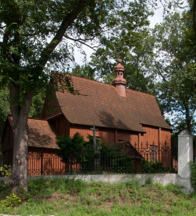 Zdjęcie nr 1: Kościół usytuowany na wzniesieniu, w otoczeniu bogatego drzewostanu. Orientowany, jednonawowy, z zamkniętym trójbocznie prezbiterium i przylegającą od północy zakrystią. Od południa i zachodu do korpusu przylegają kruchty. Nawa na planie prostokąta, szersza od prezbiterium. Wewnątrz pozorne sklepienie kolebkowe, w nawie z płaskimi odcinkami stropowymi po bokach, podpartymi na profilowanych słupach ustawionych na cokołach. Ściany opięte pilastrami naśladującymi formę słupów. Prezbiterium od korpusu oddziela łuk tęczowy, półkolisty, profilowany, ozdobiony ornamentem sznurowym, wsparty na profilowanych kroksztynach dekorowanych drewnianymi rozetami. Od zachodu parapet chóru muzycznego na planie prostokąta oparty na dwóch wielobocznych słupach, front dekorowany motywem w formie powtarzających się zwróconych do siebie par esownic i napisem „MIŁOŚCIĄ WIECZNĄ UMIŁOWAŁEM CIĘ” rozdzielonym wizerunkiem pelikana karmiącego młode własną krwią. Od południa schody prowadzące na chór z analogiczną dekoracją. Fasada prosta, zamknięta daszkiem krytym gontem, zwieńczona trójkątnie. Korpus z trzech stron wzmocniony belkami. Od strony północnej i południowej po dwa okna prostokątne zwieńczone niewielkimi blaszanymi daszkami. Prezbiterium od strony południowej opięte dwoma belkami, z prostokątnym oknem. Na ścianie wschodniej, na zewnątrz, kapliczka z ceramicznym przedstawieniem Matki Boskiej z Dzieciątkiem. W ścianach wschodniej i północnej zakrystii po jednym prostokątnym oknie. Dach dwuspadowy, o jednej kalenicy, od wschodu odzwierciedla układ ścian prezbiterium. Kruchty przykryte dachami dwuspadowymi. Dachy kryte gontem. Na przecięciu korpusu z prezbiterium blaszana, ośmioboczna wieżyczka na sygnaturkę zwieńczona hełmem baniastym ze spłaszczoną kulą z krzyżem. 