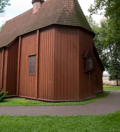 Zdjęcie nr 8: Kościół usytuowany na wzniesieniu, w otoczeniu bogatego drzewostanu. Orientowany, jednonawowy, z zamkniętym trójbocznie prezbiterium i przylegającą od północy zakrystią. Od południa i zachodu do korpusu przylegają kruchty. Nawa na planie prostokąta, szersza od prezbiterium. Wewnątrz pozorne sklepienie kolebkowe, w nawie z płaskimi odcinkami stropowymi po bokach, podpartymi na profilowanych słupach ustawionych na cokołach. Ściany opięte pilastrami naśladującymi formę słupów. Prezbiterium od korpusu oddziela łuk tęczowy, półkolisty, profilowany, ozdobiony ornamentem sznurowym, wsparty na profilowanych kroksztynach dekorowanych drewnianymi rozetami. Od zachodu parapet chóru muzycznego na planie prostokąta oparty na dwóch wielobocznych słupach, front dekorowany motywem w formie powtarzających się zwróconych do siebie par esownic i napisem „MIŁOŚCIĄ WIECZNĄ UMIŁOWAŁEM CIĘ” rozdzielonym wizerunkiem pelikana karmiącego młode własną krwią. Od południa schody prowadzące na chór z analogiczną dekoracją. Fasada prosta, zamknięta daszkiem krytym gontem, zwieńczona trójkątnie. Korpus z trzech stron wzmocniony belkami. Od strony północnej i południowej po dwa okna prostokątne zwieńczone niewielkimi blaszanymi daszkami. Prezbiterium od strony południowej opięte dwoma belkami, z prostokątnym oknem. Na ścianie wschodniej, na zewnątrz, kapliczka z ceramicznym przedstawieniem Matki Boskiej z Dzieciątkiem. W ścianach wschodniej i północnej zakrystii po jednym prostokątnym oknie. Dach dwuspadowy, o jednej kalenicy, od wschodu odzwierciedla układ ścian prezbiterium. Kruchty przykryte dachami dwuspadowymi. Dachy kryte gontem. Na przecięciu korpusu z prezbiterium blaszana, ośmioboczna wieżyczka na sygnaturkę zwieńczona hełmem baniastym ze spłaszczoną kulą z krzyżem. 