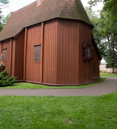 Zdjęcie nr 7: Kościół usytuowany na wzniesieniu, w otoczeniu bogatego drzewostanu. Orientowany, jednonawowy, z zamkniętym trójbocznie prezbiterium i przylegającą od północy zakrystią. Od południa i zachodu do korpusu przylegają kruchty. Nawa na planie prostokąta, szersza od prezbiterium. Wewnątrz pozorne sklepienie kolebkowe, w nawie z płaskimi odcinkami stropowymi po bokach, podpartymi na profilowanych słupach ustawionych na cokołach. Ściany opięte pilastrami naśladującymi formę słupów. Prezbiterium od korpusu oddziela łuk tęczowy, półkolisty, profilowany, ozdobiony ornamentem sznurowym, wsparty na profilowanych kroksztynach dekorowanych drewnianymi rozetami. Od zachodu parapet chóru muzycznego na planie prostokąta oparty na dwóch wielobocznych słupach, front dekorowany motywem w formie powtarzających się zwróconych do siebie par esownic i napisem „MIŁOŚCIĄ WIECZNĄ UMIŁOWAŁEM CIĘ” rozdzielonym wizerunkiem pelikana karmiącego młode własną krwią. Od południa schody prowadzące na chór z analogiczną dekoracją. Fasada prosta, zamknięta daszkiem krytym gontem, zwieńczona trójkątnie. Korpus z trzech stron wzmocniony belkami. Od strony północnej i południowej po dwa okna prostokątne zwieńczone niewielkimi blaszanymi daszkami. Prezbiterium od strony południowej opięte dwoma belkami, z prostokątnym oknem. Na ścianie wschodniej, na zewnątrz, kapliczka z ceramicznym przedstawieniem Matki Boskiej z Dzieciątkiem. W ścianach wschodniej i północnej zakrystii po jednym prostokątnym oknie. Dach dwuspadowy, o jednej kalenicy, od wschodu odzwierciedla układ ścian prezbiterium. Kruchty przykryte dachami dwuspadowymi. Dachy kryte gontem. Na przecięciu korpusu z prezbiterium blaszana, ośmioboczna wieżyczka na sygnaturkę zwieńczona hełmem baniastym ze spłaszczoną kulą z krzyżem. 