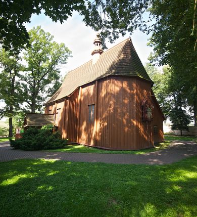 Zdjęcie nr 5: Kościół usytuowany na wzniesieniu, w otoczeniu bogatego drzewostanu. Orientowany, jednonawowy, z zamkniętym trójbocznie prezbiterium i przylegającą od północy zakrystią. Od południa i zachodu do korpusu przylegają kruchty. Nawa na planie prostokąta, szersza od prezbiterium. Wewnątrz pozorne sklepienie kolebkowe, w nawie z płaskimi odcinkami stropowymi po bokach, podpartymi na profilowanych słupach ustawionych na cokołach. Ściany opięte pilastrami naśladującymi formę słupów. Prezbiterium od korpusu oddziela łuk tęczowy, półkolisty, profilowany, ozdobiony ornamentem sznurowym, wsparty na profilowanych kroksztynach dekorowanych drewnianymi rozetami. Od zachodu parapet chóru muzycznego na planie prostokąta oparty na dwóch wielobocznych słupach, front dekorowany motywem w formie powtarzających się zwróconych do siebie par esownic i napisem „MIŁOŚCIĄ WIECZNĄ UMIŁOWAŁEM CIĘ” rozdzielonym wizerunkiem pelikana karmiącego młode własną krwią. Od południa schody prowadzące na chór z analogiczną dekoracją. Fasada prosta, zamknięta daszkiem krytym gontem, zwieńczona trójkątnie. Korpus z trzech stron wzmocniony belkami. Od strony północnej i południowej po dwa okna prostokątne zwieńczone niewielkimi blaszanymi daszkami. Prezbiterium od strony południowej opięte dwoma belkami, z prostokątnym oknem. Na ścianie wschodniej, na zewnątrz, kapliczka z ceramicznym przedstawieniem Matki Boskiej z Dzieciątkiem. W ścianach wschodniej i północnej zakrystii po jednym prostokątnym oknie. Dach dwuspadowy, o jednej kalenicy, od wschodu odzwierciedla układ ścian prezbiterium. Kruchty przykryte dachami dwuspadowymi. Dachy kryte gontem. Na przecięciu korpusu z prezbiterium blaszana, ośmioboczna wieżyczka na sygnaturkę zwieńczona hełmem baniastym ze spłaszczoną kulą z krzyżem. 