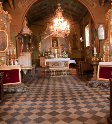 Zdjęcie nr 22: Kościół usytuowany na wzniesieniu, w otoczeniu bogatego drzewostanu. Orientowany, jednonawowy, z zamkniętym trójbocznie prezbiterium i przylegającą od północy zakrystią. Od południa i zachodu do korpusu przylegają kruchty. Nawa na planie prostokąta, szersza od prezbiterium. Wewnątrz pozorne sklepienie kolebkowe, w nawie z płaskimi odcinkami stropowymi po bokach, podpartymi na profilowanych słupach ustawionych na cokołach. Ściany opięte pilastrami naśladującymi formę słupów. Prezbiterium od korpusu oddziela łuk tęczowy, półkolisty, profilowany, ozdobiony ornamentem sznurowym, wsparty na profilowanych kroksztynach dekorowanych drewnianymi rozetami. Od zachodu parapet chóru muzycznego na planie prostokąta oparty na dwóch wielobocznych słupach, front dekorowany motywem w formie powtarzających się zwróconych do siebie par esownic i napisem „MIŁOŚCIĄ WIECZNĄ UMIŁOWAŁEM CIĘ” rozdzielonym wizerunkiem pelikana karmiącego młode własną krwią. Od południa schody prowadzące na chór z analogiczną dekoracją. Fasada prosta, zamknięta daszkiem krytym gontem, zwieńczona trójkątnie. Korpus z trzech stron wzmocniony belkami. Od strony północnej i południowej po dwa okna prostokątne zwieńczone niewielkimi blaszanymi daszkami. Prezbiterium od strony południowej opięte dwoma belkami, z prostokątnym oknem. Na ścianie wschodniej, na zewnątrz, kapliczka z ceramicznym przedstawieniem Matki Boskiej z Dzieciątkiem. W ścianach wschodniej i północnej zakrystii po jednym prostokątnym oknie. Dach dwuspadowy, o jednej kalenicy, od wschodu odzwierciedla układ ścian prezbiterium. Kruchty przykryte dachami dwuspadowymi. Dachy kryte gontem. Na przecięciu korpusu z prezbiterium blaszana, ośmioboczna wieżyczka na sygnaturkę zwieńczona hełmem baniastym ze spłaszczoną kulą z krzyżem. 