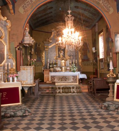 Zdjęcie nr 23: Kościół usytuowany na wzniesieniu, w otoczeniu bogatego drzewostanu. Orientowany, jednonawowy, z zamkniętym trójbocznie prezbiterium i przylegającą od północy zakrystią. Od południa i zachodu do korpusu przylegają kruchty. Nawa na planie prostokąta, szersza od prezbiterium. Wewnątrz pozorne sklepienie kolebkowe, w nawie z płaskimi odcinkami stropowymi po bokach, podpartymi na profilowanych słupach ustawionych na cokołach. Ściany opięte pilastrami naśladującymi formę słupów. Prezbiterium od korpusu oddziela łuk tęczowy, półkolisty, profilowany, ozdobiony ornamentem sznurowym, wsparty na profilowanych kroksztynach dekorowanych drewnianymi rozetami. Od zachodu parapet chóru muzycznego na planie prostokąta oparty na dwóch wielobocznych słupach, front dekorowany motywem w formie powtarzających się zwróconych do siebie par esownic i napisem „MIŁOŚCIĄ WIECZNĄ UMIŁOWAŁEM CIĘ” rozdzielonym wizerunkiem pelikana karmiącego młode własną krwią. Od południa schody prowadzące na chór z analogiczną dekoracją. Fasada prosta, zamknięta daszkiem krytym gontem, zwieńczona trójkątnie. Korpus z trzech stron wzmocniony belkami. Od strony północnej i południowej po dwa okna prostokątne zwieńczone niewielkimi blaszanymi daszkami. Prezbiterium od strony południowej opięte dwoma belkami, z prostokątnym oknem. Na ścianie wschodniej, na zewnątrz, kapliczka z ceramicznym przedstawieniem Matki Boskiej z Dzieciątkiem. W ścianach wschodniej i północnej zakrystii po jednym prostokątnym oknie. Dach dwuspadowy, o jednej kalenicy, od wschodu odzwierciedla układ ścian prezbiterium. Kruchty przykryte dachami dwuspadowymi. Dachy kryte gontem. Na przecięciu korpusu z prezbiterium blaszana, ośmioboczna wieżyczka na sygnaturkę zwieńczona hełmem baniastym ze spłaszczoną kulą z krzyżem. 