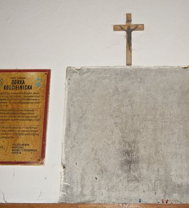 Zdjęcie nr 20: Kościół usytuowany na wzniesieniu, w otoczeniu bogatego drzewostanu. Orientowany, jednonawowy, z zamkniętym trójbocznie prezbiterium i przylegającą od północy zakrystią. Od południa i zachodu do korpusu przylegają kruchty. Nawa na planie prostokąta, szersza od prezbiterium. Wewnątrz pozorne sklepienie kolebkowe, w nawie z płaskimi odcinkami stropowymi po bokach, podpartymi na profilowanych słupach ustawionych na cokołach. Ściany opięte pilastrami naśladującymi formę słupów. Prezbiterium od korpusu oddziela łuk tęczowy, półkolisty, profilowany, ozdobiony ornamentem sznurowym, wsparty na profilowanych kroksztynach dekorowanych drewnianymi rozetami. Od zachodu parapet chóru muzycznego na planie prostokąta oparty na dwóch wielobocznych słupach, front dekorowany motywem w formie powtarzających się zwróconych do siebie par esownic i napisem „MIŁOŚCIĄ WIECZNĄ UMIŁOWAŁEM CIĘ” rozdzielonym wizerunkiem pelikana karmiącego młode własną krwią. Od południa schody prowadzące na chór z analogiczną dekoracją. Fasada prosta, zamknięta daszkiem krytym gontem, zwieńczona trójkątnie. Korpus z trzech stron wzmocniony belkami. Od strony północnej i południowej po dwa okna prostokątne zwieńczone niewielkimi blaszanymi daszkami. Prezbiterium od strony południowej opięte dwoma belkami, z prostokątnym oknem. Na ścianie wschodniej, na zewnątrz, kapliczka z ceramicznym przedstawieniem Matki Boskiej z Dzieciątkiem. W ścianach wschodniej i północnej zakrystii po jednym prostokątnym oknie. Dach dwuspadowy, o jednej kalenicy, od wschodu odzwierciedla układ ścian prezbiterium. Kruchty przykryte dachami dwuspadowymi. Dachy kryte gontem. Na przecięciu korpusu z prezbiterium blaszana, ośmioboczna wieżyczka na sygnaturkę zwieńczona hełmem baniastym ze spłaszczoną kulą z krzyżem. 