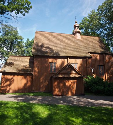 Zdjęcie nr 26: Kościół usytuowany na wzniesieniu, w otoczeniu bogatego drzewostanu. Orientowany, jednonawowy, z zamkniętym trójbocznie prezbiterium i przylegającą od północy zakrystią. Od południa i zachodu do korpusu przylegają kruchty. Nawa na planie prostokąta, szersza od prezbiterium. Wewnątrz pozorne sklepienie kolebkowe, w nawie z płaskimi odcinkami stropowymi po bokach, podpartymi na profilowanych słupach ustawionych na cokołach. Ściany opięte pilastrami naśladującymi formę słupów. Prezbiterium od korpusu oddziela łuk tęczowy, półkolisty, profilowany, ozdobiony ornamentem sznurowym, wsparty na profilowanych kroksztynach dekorowanych drewnianymi rozetami. Od zachodu parapet chóru muzycznego na planie prostokąta oparty na dwóch wielobocznych słupach, front dekorowany motywem w formie powtarzających się zwróconych do siebie par esownic i napisem „MIŁOŚCIĄ WIECZNĄ UMIŁOWAŁEM CIĘ” rozdzielonym wizerunkiem pelikana karmiącego młode własną krwią. Od południa schody prowadzące na chór z analogiczną dekoracją. Fasada prosta, zamknięta daszkiem krytym gontem, zwieńczona trójkątnie. Korpus z trzech stron wzmocniony belkami. Od strony północnej i południowej po dwa okna prostokątne zwieńczone niewielkimi blaszanymi daszkami. Prezbiterium od strony południowej opięte dwoma belkami, z prostokątnym oknem. Na ścianie wschodniej, na zewnątrz, kapliczka z ceramicznym przedstawieniem Matki Boskiej z Dzieciątkiem. W ścianach wschodniej i północnej zakrystii po jednym prostokątnym oknie. Dach dwuspadowy, o jednej kalenicy, od wschodu odzwierciedla układ ścian prezbiterium. Kruchty przykryte dachami dwuspadowymi. Dachy kryte gontem. Na przecięciu korpusu z prezbiterium blaszana, ośmioboczna wieżyczka na sygnaturkę zwieńczona hełmem baniastym ze spłaszczoną kulą z krzyżem. 