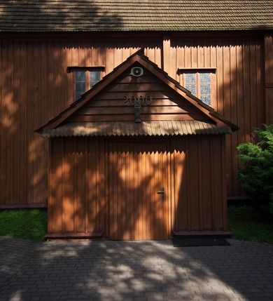 Zdjęcie nr 4: Kościół usytuowany na wzniesieniu, w otoczeniu bogatego drzewostanu. Orientowany, jednonawowy, z zamkniętym trójbocznie prezbiterium i przylegającą od północy zakrystią. Od południa i zachodu do korpusu przylegają kruchty. Nawa na planie prostokąta, szersza od prezbiterium. Wewnątrz pozorne sklepienie kolebkowe, w nawie z płaskimi odcinkami stropowymi po bokach, podpartymi na profilowanych słupach ustawionych na cokołach. Ściany opięte pilastrami naśladującymi formę słupów. Prezbiterium od korpusu oddziela łuk tęczowy, półkolisty, profilowany, ozdobiony ornamentem sznurowym, wsparty na profilowanych kroksztynach dekorowanych drewnianymi rozetami. Od zachodu parapet chóru muzycznego na planie prostokąta oparty na dwóch wielobocznych słupach, front dekorowany motywem w formie powtarzających się zwróconych do siebie par esownic i napisem „MIŁOŚCIĄ WIECZNĄ UMIŁOWAŁEM CIĘ” rozdzielonym wizerunkiem pelikana karmiącego młode własną krwią. Od południa schody prowadzące na chór z analogiczną dekoracją. Fasada prosta, zamknięta daszkiem krytym gontem, zwieńczona trójkątnie. Korpus z trzech stron wzmocniony belkami. Od strony północnej i południowej po dwa okna prostokątne zwieńczone niewielkimi blaszanymi daszkami. Prezbiterium od strony południowej opięte dwoma belkami, z prostokątnym oknem. Na ścianie wschodniej, na zewnątrz, kapliczka z ceramicznym przedstawieniem Matki Boskiej z Dzieciątkiem. W ścianach wschodniej i północnej zakrystii po jednym prostokątnym oknie. Dach dwuspadowy, o jednej kalenicy, od wschodu odzwierciedla układ ścian prezbiterium. Kruchty przykryte dachami dwuspadowymi. Dachy kryte gontem. Na przecięciu korpusu z prezbiterium blaszana, ośmioboczna wieżyczka na sygnaturkę zwieńczona hełmem baniastym ze spłaszczoną kulą z krzyżem. 