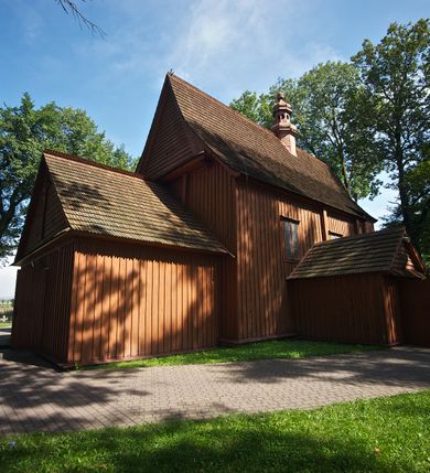 Zdjęcie nr 27: Kościół usytuowany na wzniesieniu, w otoczeniu bogatego drzewostanu. Orientowany, jednonawowy, z zamkniętym trójbocznie prezbiterium i przylegającą od północy zakrystią. Od południa i zachodu do korpusu przylegają kruchty. Nawa na planie prostokąta, szersza od prezbiterium. Wewnątrz pozorne sklepienie kolebkowe, w nawie z płaskimi odcinkami stropowymi po bokach, podpartymi na profilowanych słupach ustawionych na cokołach. Ściany opięte pilastrami naśladującymi formę słupów. Prezbiterium od korpusu oddziela łuk tęczowy, półkolisty, profilowany, ozdobiony ornamentem sznurowym, wsparty na profilowanych kroksztynach dekorowanych drewnianymi rozetami. Od zachodu parapet chóru muzycznego na planie prostokąta oparty na dwóch wielobocznych słupach, front dekorowany motywem w formie powtarzających się zwróconych do siebie par esownic i napisem „MIŁOŚCIĄ WIECZNĄ UMIŁOWAŁEM CIĘ” rozdzielonym wizerunkiem pelikana karmiącego młode własną krwią. Od południa schody prowadzące na chór z analogiczną dekoracją. Fasada prosta, zamknięta daszkiem krytym gontem, zwieńczona trójkątnie. Korpus z trzech stron wzmocniony belkami. Od strony północnej i południowej po dwa okna prostokątne zwieńczone niewielkimi blaszanymi daszkami. Prezbiterium od strony południowej opięte dwoma belkami, z prostokątnym oknem. Na ścianie wschodniej, na zewnątrz, kapliczka z ceramicznym przedstawieniem Matki Boskiej z Dzieciątkiem. W ścianach wschodniej i północnej zakrystii po jednym prostokątnym oknie. Dach dwuspadowy, o jednej kalenicy, od wschodu odzwierciedla układ ścian prezbiterium. Kruchty przykryte dachami dwuspadowymi. Dachy kryte gontem. Na przecięciu korpusu z prezbiterium blaszana, ośmioboczna wieżyczka na sygnaturkę zwieńczona hełmem baniastym ze spłaszczoną kulą z krzyżem. 