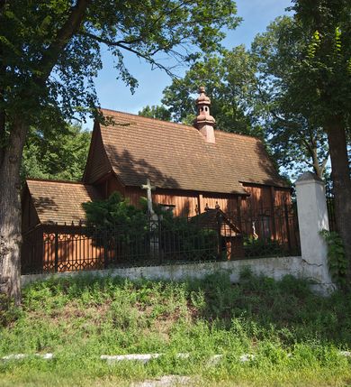 Zdjęcie nr 24: Kościół usytuowany na wzniesieniu, w otoczeniu bogatego drzewostanu. Orientowany, jednonawowy, z zamkniętym trójbocznie prezbiterium i przylegającą od północy zakrystią. Od południa i zachodu do korpusu przylegają kruchty. Nawa na planie prostokąta, szersza od prezbiterium. Wewnątrz pozorne sklepienie kolebkowe, w nawie z płaskimi odcinkami stropowymi po bokach, podpartymi na profilowanych słupach ustawionych na cokołach. Ściany opięte pilastrami naśladującymi formę słupów. Prezbiterium od korpusu oddziela łuk tęczowy, półkolisty, profilowany, ozdobiony ornamentem sznurowym, wsparty na profilowanych kroksztynach dekorowanych drewnianymi rozetami. Od zachodu parapet chóru muzycznego na planie prostokąta oparty na dwóch wielobocznych słupach, front dekorowany motywem w formie powtarzających się zwróconych do siebie par esownic i napisem „MIŁOŚCIĄ WIECZNĄ UMIŁOWAŁEM CIĘ” rozdzielonym wizerunkiem pelikana karmiącego młode własną krwią. Od południa schody prowadzące na chór z analogiczną dekoracją. Fasada prosta, zamknięta daszkiem krytym gontem, zwieńczona trójkątnie. Korpus z trzech stron wzmocniony belkami. Od strony północnej i południowej po dwa okna prostokątne zwieńczone niewielkimi blaszanymi daszkami. Prezbiterium od strony południowej opięte dwoma belkami, z prostokątnym oknem. Na ścianie wschodniej, na zewnątrz, kapliczka z ceramicznym przedstawieniem Matki Boskiej z Dzieciątkiem. W ścianach wschodniej i północnej zakrystii po jednym prostokątnym oknie. Dach dwuspadowy, o jednej kalenicy, od wschodu odzwierciedla układ ścian prezbiterium. Kruchty przykryte dachami dwuspadowymi. Dachy kryte gontem. Na przecięciu korpusu z prezbiterium blaszana, ośmioboczna wieżyczka na sygnaturkę zwieńczona hełmem baniastym ze spłaszczoną kulą z krzyżem. 