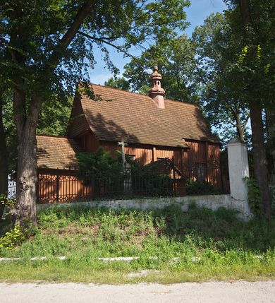 Zdjęcie nr 13: Kościół usytuowany na wzniesieniu, w otoczeniu bogatego drzewostanu. Orientowany, jednonawowy, z zamkniętym trójbocznie prezbiterium i przylegającą od północy zakrystią. Od południa i zachodu do korpusu przylegają kruchty. Nawa na planie prostokąta, szersza od prezbiterium. Wewnątrz pozorne sklepienie kolebkowe, w nawie z płaskimi odcinkami stropowymi po bokach, podpartymi na profilowanych słupach ustawionych na cokołach. Ściany opięte pilastrami naśladującymi formę słupów. Prezbiterium od korpusu oddziela łuk tęczowy, półkolisty, profilowany, ozdobiony ornamentem sznurowym, wsparty na profilowanych kroksztynach dekorowanych drewnianymi rozetami. Od zachodu parapet chóru muzycznego na planie prostokąta oparty na dwóch wielobocznych słupach, front dekorowany motywem w formie powtarzających się zwróconych do siebie par esownic i napisem „MIŁOŚCIĄ WIECZNĄ UMIŁOWAŁEM CIĘ” rozdzielonym wizerunkiem pelikana karmiącego młode własną krwią. Od południa schody prowadzące na chór z analogiczną dekoracją. Fasada prosta, zamknięta daszkiem krytym gontem, zwieńczona trójkątnie. Korpus z trzech stron wzmocniony belkami. Od strony północnej i południowej po dwa okna prostokątne zwieńczone niewielkimi blaszanymi daszkami. Prezbiterium od strony południowej opięte dwoma belkami, z prostokątnym oknem. Na ścianie wschodniej, na zewnątrz, kapliczka z ceramicznym przedstawieniem Matki Boskiej z Dzieciątkiem. W ścianach wschodniej i północnej zakrystii po jednym prostokątnym oknie. Dach dwuspadowy, o jednej kalenicy, od wschodu odzwierciedla układ ścian prezbiterium. Kruchty przykryte dachami dwuspadowymi. Dachy kryte gontem. Na przecięciu korpusu z prezbiterium blaszana, ośmioboczna wieżyczka na sygnaturkę zwieńczona hełmem baniastym ze spłaszczoną kulą z krzyżem. 