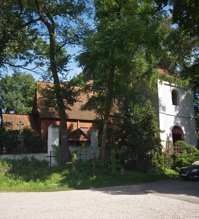 Zdjęcie nr 2: Kościół usytuowany na wzniesieniu, w otoczeniu bogatego drzewostanu. Orientowany, jednonawowy, z zamkniętym trójbocznie prezbiterium i przylegającą od północy zakrystią. Od południa i zachodu do korpusu przylegają kruchty. Nawa na planie prostokąta, szersza od prezbiterium. Wewnątrz pozorne sklepienie kolebkowe, w nawie z płaskimi odcinkami stropowymi po bokach, podpartymi na profilowanych słupach ustawionych na cokołach. Ściany opięte pilastrami naśladującymi formę słupów. Prezbiterium od korpusu oddziela łuk tęczowy, półkolisty, profilowany, ozdobiony ornamentem sznurowym, wsparty na profilowanych kroksztynach dekorowanych drewnianymi rozetami. Od zachodu parapet chóru muzycznego na planie prostokąta oparty na dwóch wielobocznych słupach, front dekorowany motywem w formie powtarzających się zwróconych do siebie par esownic i napisem „MIŁOŚCIĄ WIECZNĄ UMIŁOWAŁEM CIĘ” rozdzielonym wizerunkiem pelikana karmiącego młode własną krwią. Od południa schody prowadzące na chór z analogiczną dekoracją. Fasada prosta, zamknięta daszkiem krytym gontem, zwieńczona trójkątnie. Korpus z trzech stron wzmocniony belkami. Od strony północnej i południowej po dwa okna prostokątne zwieńczone niewielkimi blaszanymi daszkami. Prezbiterium od strony południowej opięte dwoma belkami, z prostokątnym oknem. Na ścianie wschodniej, na zewnątrz, kapliczka z ceramicznym przedstawieniem Matki Boskiej z Dzieciątkiem. W ścianach wschodniej i północnej zakrystii po jednym prostokątnym oknie. Dach dwuspadowy, o jednej kalenicy, od wschodu odzwierciedla układ ścian prezbiterium. Kruchty przykryte dachami dwuspadowymi. Dachy kryte gontem. Na przecięciu korpusu z prezbiterium blaszana, ośmioboczna wieżyczka na sygnaturkę zwieńczona hełmem baniastym ze spłaszczoną kulą z krzyżem. 