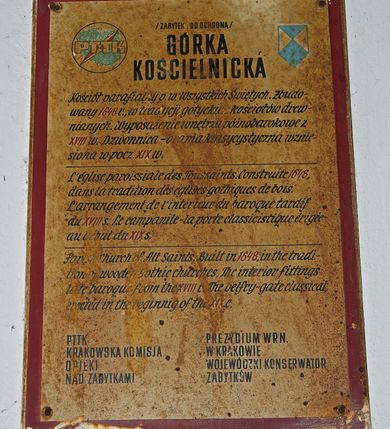 Zdjęcie nr 21: Kościół usytuowany na wzniesieniu, w otoczeniu bogatego drzewostanu. Orientowany, jednonawowy, z zamkniętym trójbocznie prezbiterium i przylegającą od północy zakrystią. Od południa i zachodu do korpusu przylegają kruchty. Nawa na planie prostokąta, szersza od prezbiterium. Wewnątrz pozorne sklepienie kolebkowe, w nawie z płaskimi odcinkami stropowymi po bokach, podpartymi na profilowanych słupach ustawionych na cokołach. Ściany opięte pilastrami naśladującymi formę słupów. Prezbiterium od korpusu oddziela łuk tęczowy, półkolisty, profilowany, ozdobiony ornamentem sznurowym, wsparty na profilowanych kroksztynach dekorowanych drewnianymi rozetami. Od zachodu parapet chóru muzycznego na planie prostokąta oparty na dwóch wielobocznych słupach, front dekorowany motywem w formie powtarzających się zwróconych do siebie par esownic i napisem „MIŁOŚCIĄ WIECZNĄ UMIŁOWAŁEM CIĘ” rozdzielonym wizerunkiem pelikana karmiącego młode własną krwią. Od południa schody prowadzące na chór z analogiczną dekoracją. Fasada prosta, zamknięta daszkiem krytym gontem, zwieńczona trójkątnie. Korpus z trzech stron wzmocniony belkami. Od strony północnej i południowej po dwa okna prostokątne zwieńczone niewielkimi blaszanymi daszkami. Prezbiterium od strony południowej opięte dwoma belkami, z prostokątnym oknem. Na ścianie wschodniej, na zewnątrz, kapliczka z ceramicznym przedstawieniem Matki Boskiej z Dzieciątkiem. W ścianach wschodniej i północnej zakrystii po jednym prostokątnym oknie. Dach dwuspadowy, o jednej kalenicy, od wschodu odzwierciedla układ ścian prezbiterium. Kruchty przykryte dachami dwuspadowymi. Dachy kryte gontem. Na przecięciu korpusu z prezbiterium blaszana, ośmioboczna wieżyczka na sygnaturkę zwieńczona hełmem baniastym ze spłaszczoną kulą z krzyżem. 