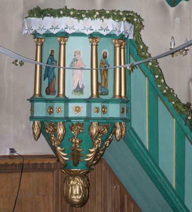 Zdjęcie nr 1: Ambona wisząca, przyścienna, z wejściem od prawej strony w postaci jednobiegowych schodów z balustradą. Kosz na planie koła otoczony kolumnami korynckimi posadowionymi na cokołach i podtrzymującymi belkowanie wyłamane na ich linii. Podwieszenie w formie stożka zakończonego dużą szyszką, dekorowanego plakietami skomponowanymi z elementów floralnych oraz  podwieszonymi szyszkami. Ścianki dekorowane malowanymi przedstawieniami  czterech ewangelistów. 
Baldachim powtarza pomniejszony plan kosza; dekorowany szyszkami, w podniebieniu lampa w kształcie szyszki. W jego zwieńczeniu umieszczono figurą Chrystusa Zbawiciela Świata (Salvator Mundi) na dwustopniowym cokole. Chrystus stoi w kontrapoście, z prawą nogą ugiętą w kolanie i wspartą na kamieniu. Lewą ręką podtrzymuje kulę ziemską zwieńczoną krzyżem, prawą unosi do góry, czyniąc gest błogosławieństwa. Spogląda do góry, ma ciemną brodę i falowane włosy spływające na ramiona i plecy. Z trzech stron głowy odchodzą wiązki promieni. Chrystus ubrany jest w długą, złotą suknię oraz tego samego koloru płaszcz owinięty wokół prawego biodra i przerzucony przez lewą rękę. 