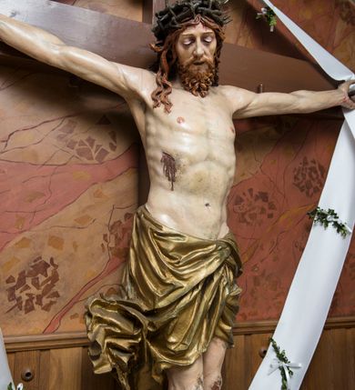 Zdjęcie nr 3: Pełnoplastyczna, polichromowana figura Chrystusa przybitego do krzyża. Ciało o jasnej karnacji i drobiazgowo opracowanej anatomii. Głowa pochylona w dół, lekko w prawo. Twarz owalna o łagodnych rysach, z zamkniętymi, otoczonymi fioletowym cieniem oczami, wąskim nosie i zamkniętych, sinych ustach; z krótkim, brązowym zarostem. Włosy długie, falowane, jasnobrązowe, opadające na plecy i prawe ramię. Na głowie korona cierniowa. Ramiona rozłożone, w prawym boku rana. Nogi wyprostowane, przebite jednym gwoździem, prawa na lewej. Na biodrach krótkie, złote, silnie drapowane, przewiązane na lewym biodrze perizonium, rozwiewające się gęstymi, łamanymi fałdami na wysokości uda w lewą stronę. W środkowej partii pionowej belki biały titulus w formie zwoju z czarnym napisem „INRI”.