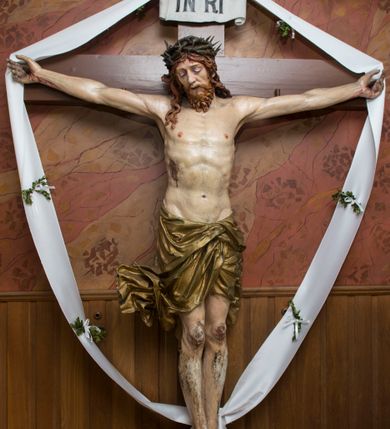 Zdjęcie nr 2: Pełnoplastyczna, polichromowana figura Chrystusa przybitego do krzyża. Ciało o jasnej karnacji i drobiazgowo opracowanej anatomii. Głowa pochylona w dół, lekko w prawo. Twarz owalna o łagodnych rysach, z zamkniętymi, otoczonymi fioletowym cieniem oczami, wąskim nosie i zamkniętych, sinych ustach; z krótkim, brązowym zarostem. Włosy długie, falowane, jasnobrązowe, opadające na plecy i prawe ramię. Na głowie korona cierniowa. Ramiona rozłożone, w prawym boku rana. Nogi wyprostowane, przebite jednym gwoździem, prawa na lewej. Na biodrach krótkie, złote, silnie drapowane, przewiązane na lewym biodrze perizonium, rozwiewające się gęstymi, łamanymi fałdami na wysokości uda w lewą stronę. W środkowej partii pionowej belki biały titulus w formie zwoju z czarnym napisem „INRI”.