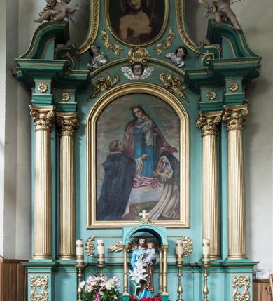 Zdjęcie nr 3: Obraz z przedstawieniem tronującej Matki Boskiej z Dzieciątkiem, przekazujących różańce klęczącym przed nimi 
 – św. Dominikowi i św. Katarzynie Sieneńskiej. Maria siedzi, spogląda delikatnie w dół. Na jej lewym kolanie stoi Dzieciątko, które podtrzymuje prawą ręką, lewą przekazując różaniec świętej. Maria ma trójkątną twarz z ciemnymi, otoczonymi cieniami oczami, wąskim nosem oraz małymi ustami. Jest ubrana w niebieską suknię oraz różowy, opadający drobnymi kaskadami na boki płaszcz, który fałduje się obficie pod jej stopami. Na głowie ma zdobioną drogimi kamieniami koronę. Dzieciątko spogląda na św. Dominika, prawą rękę wyciąga w jego kierunku w geście błogosławieństwa, lewą podaje mu różaniec. Ma okrągłą twarz, ciemne oczy, mały nos i usta, nieznacznie się uśmiecha. Jest ubrane w białą, długą sukienkę. Po lewej stronie przedstawienia klęczy św. Dominik, który spogląda na Dzieciątko i prawą ręką odbiera od niego różaniec. Ubrany jest w biały habit i czarny płaszcz zakonny, na głowie ma wyciętą tonsurę. Klęcząca po prawej stronie św. Katarzyna patrzy na podtrzymywany lewą ręką różaniec, prawą składa na piersi. Ubrana jest w biały habit i czarny płaszcz, na głowie ma koronę cierniową. Scena rozgrywa się na tle niebieskiego nieba przykrytego gęsto żółtopomarańczowymi chmurami. Między klęczącymi świętymi wyrasta z ziemi kwitnący krzew różany.